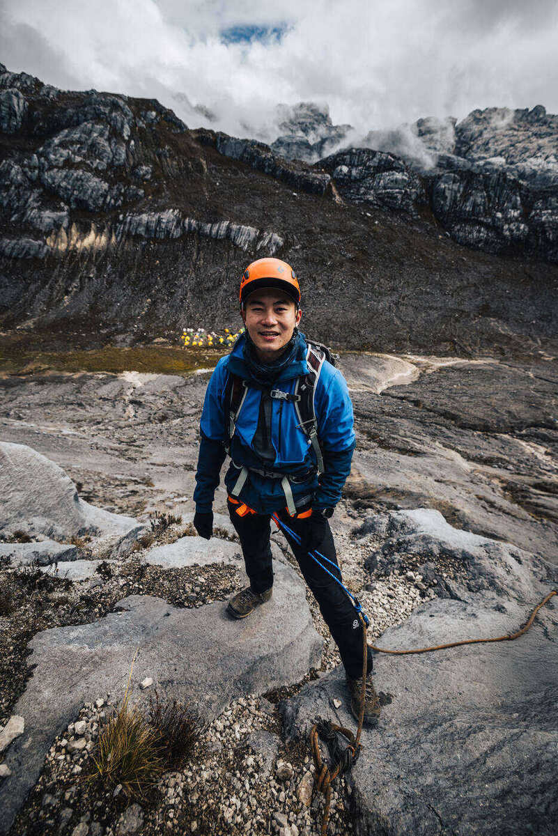 Climber in blue jacket and orange helmet with backpack posing on rocky ground with limestone slopes behind