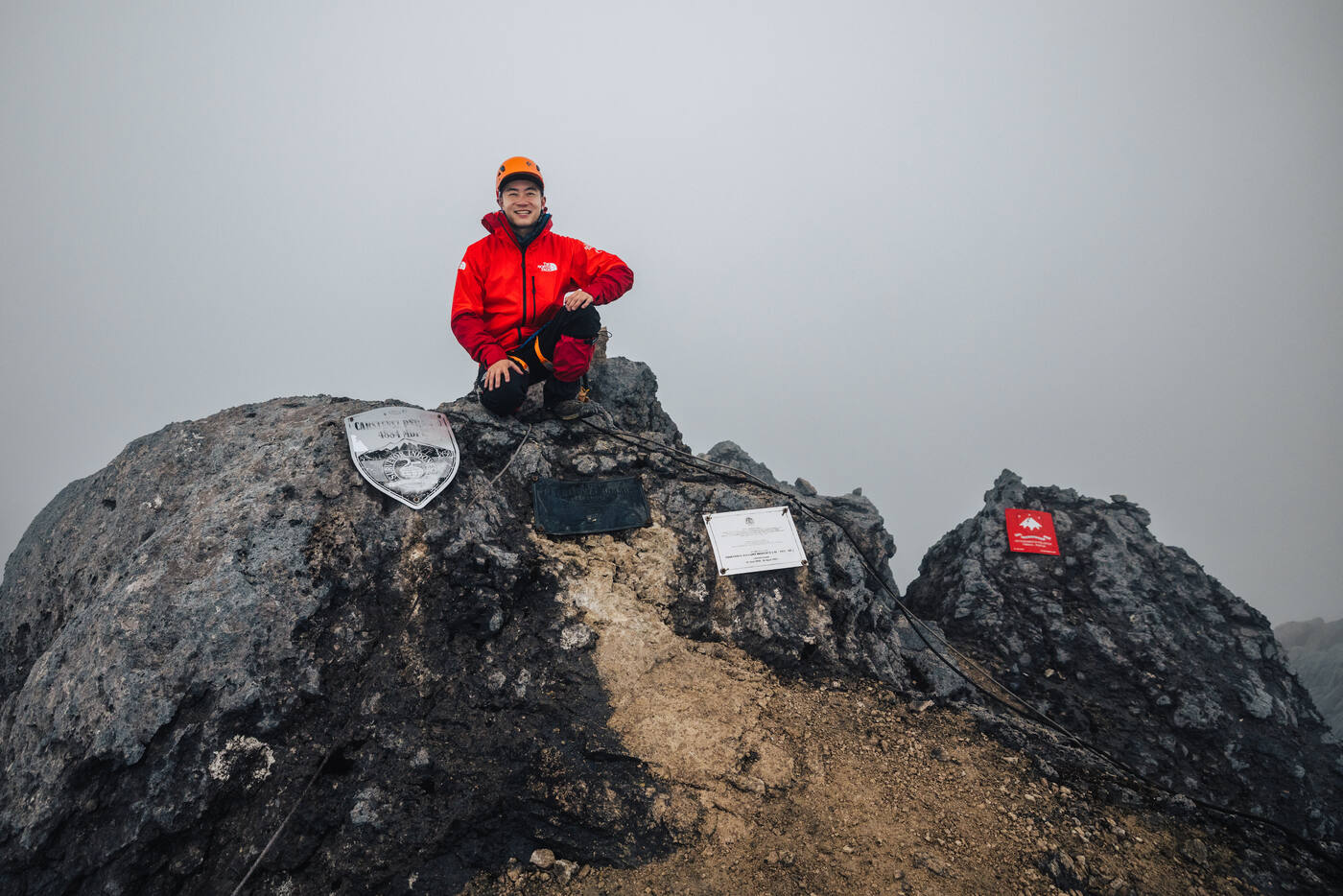 Climber in red jacket crouched on the dark summit rock above the summit plaques