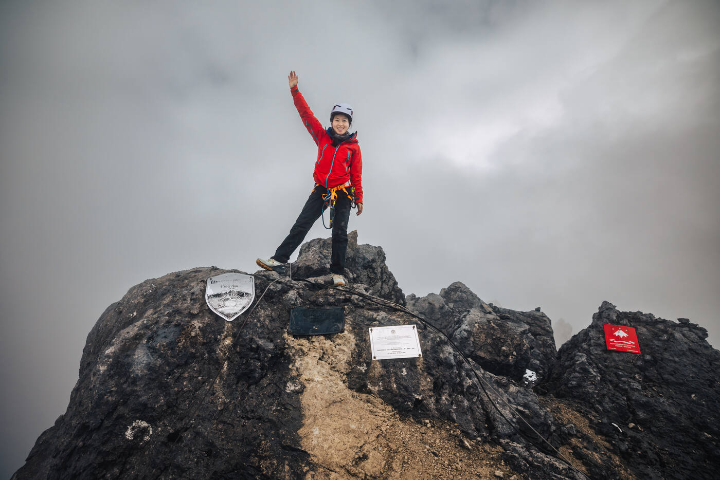 Grace standing on the summit of Carstensz Pyramid in red jacket with arm raised, summit plaques visible