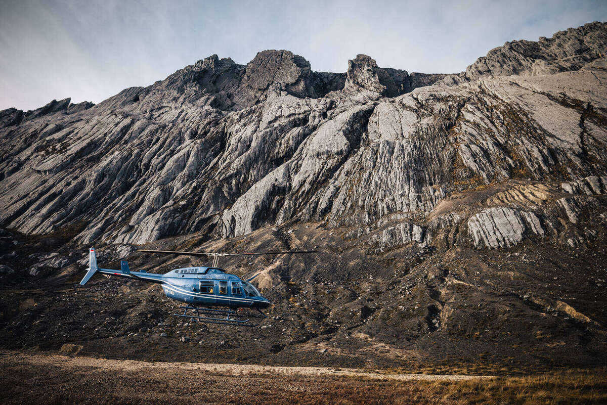 Blue helicopter parked on rocky terrain at the base of a massive limestone cliff wall