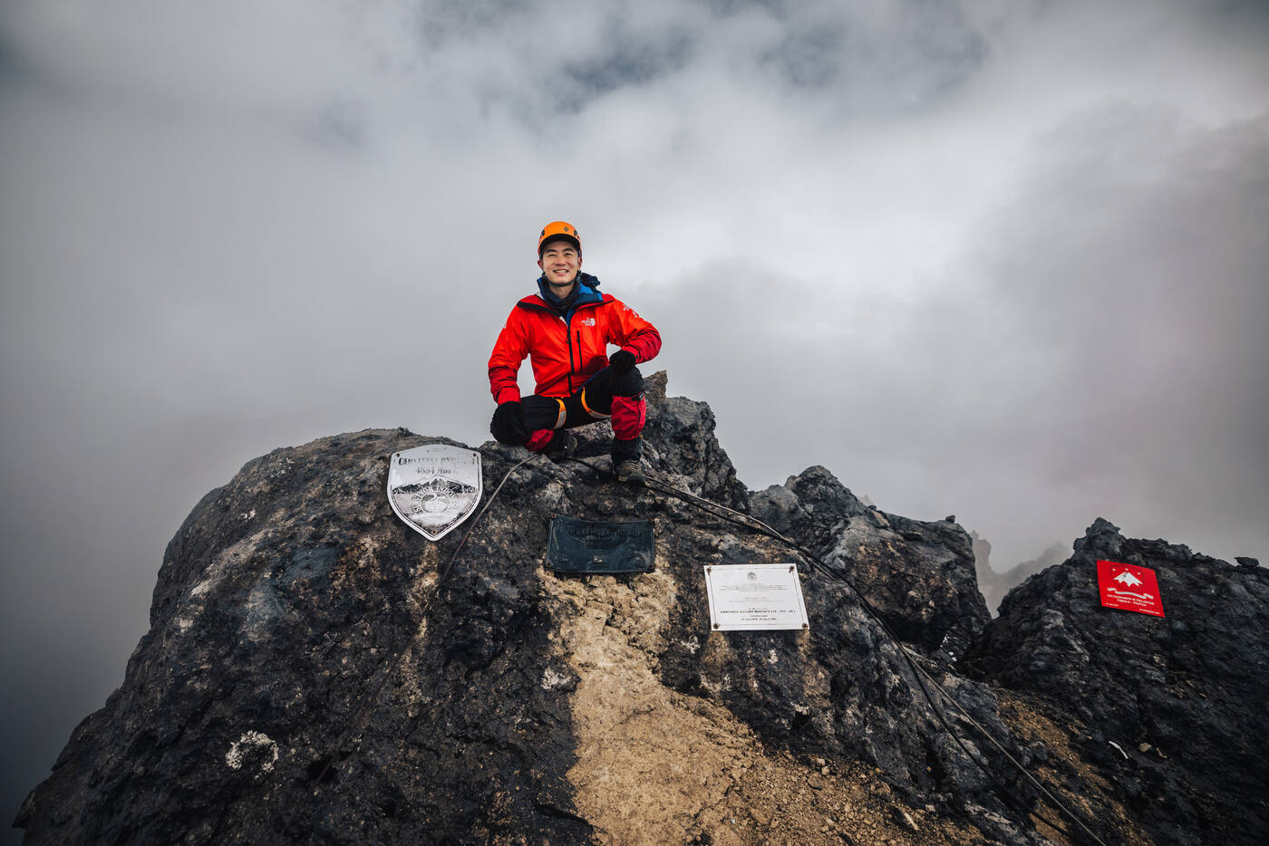 Climber in red jacket and orange helmet crouched on the summit rock beside the summit plaques, cloud behind