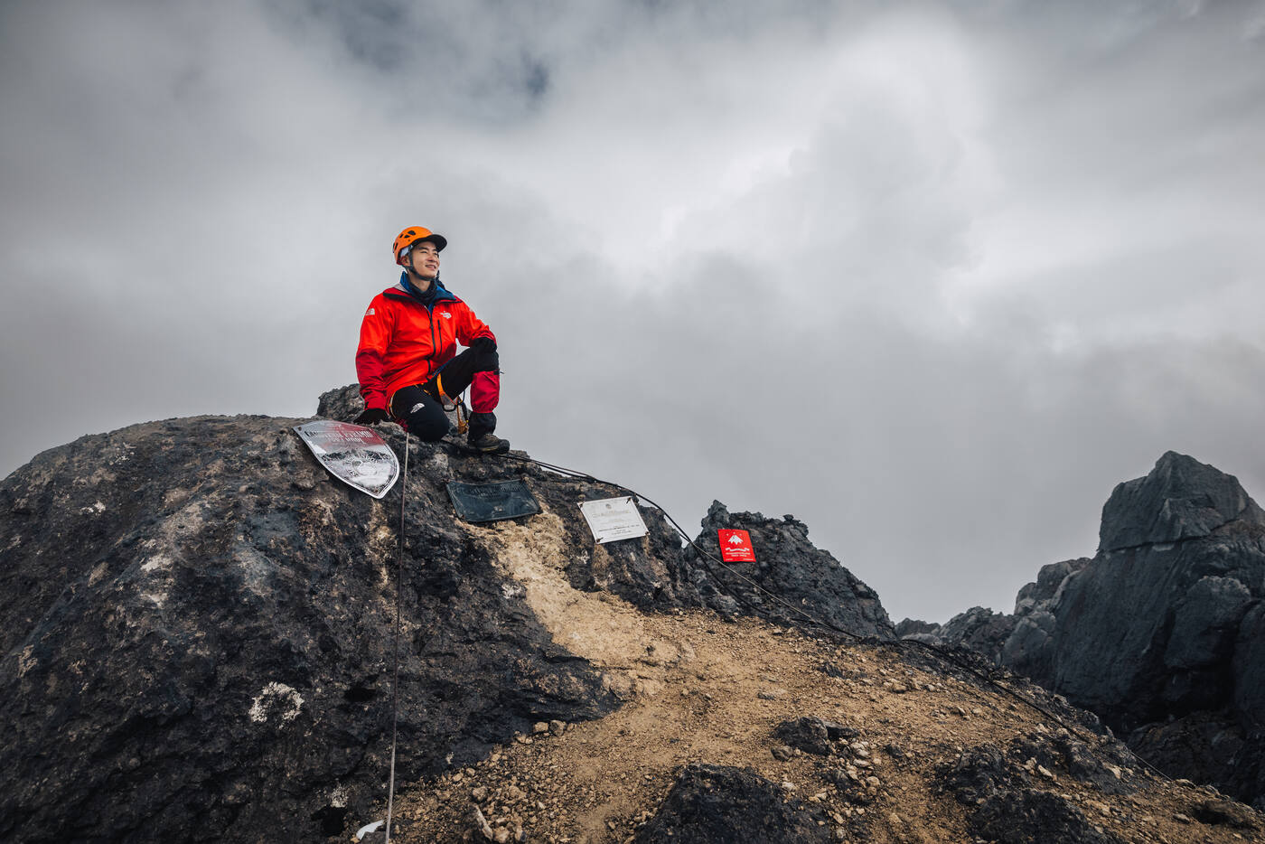 Climber in red seated on the summit rock beside the shield-shaped summit plaque, storm clouds behind