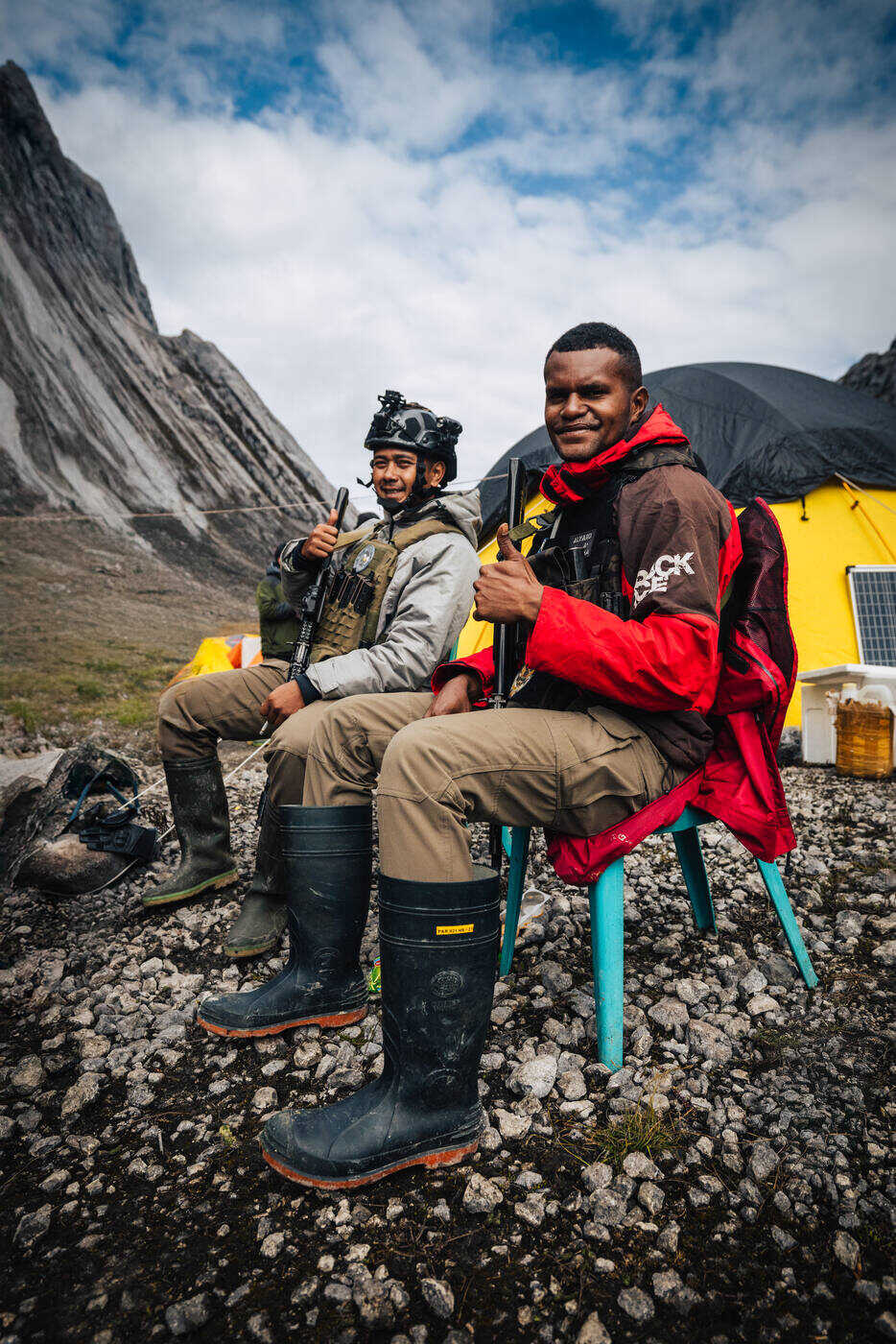 Two team members sitting on chairs at base camp with rock walls and yellow tent behind