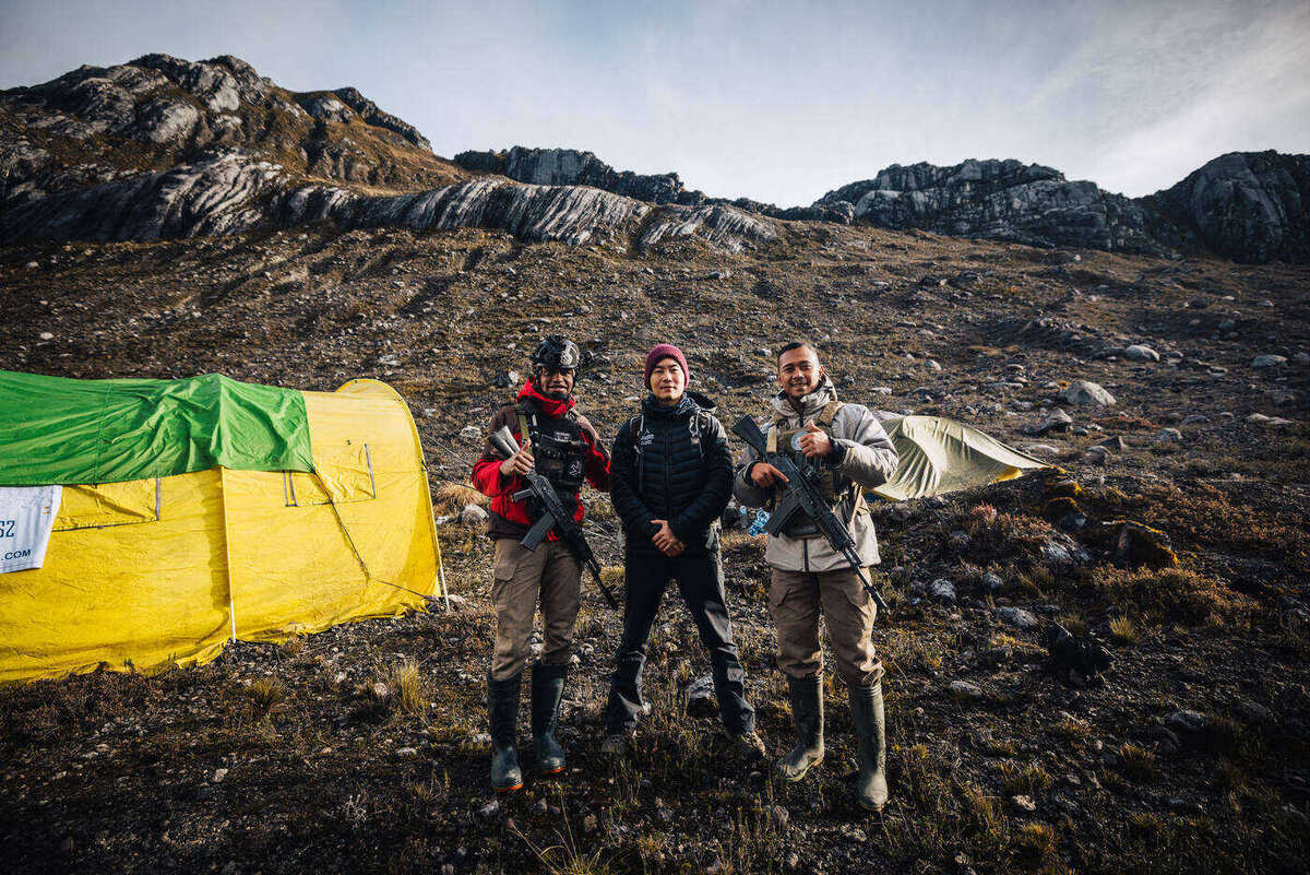 Three team members posed at base camp with yellow tent and rocky mountain valley behind
