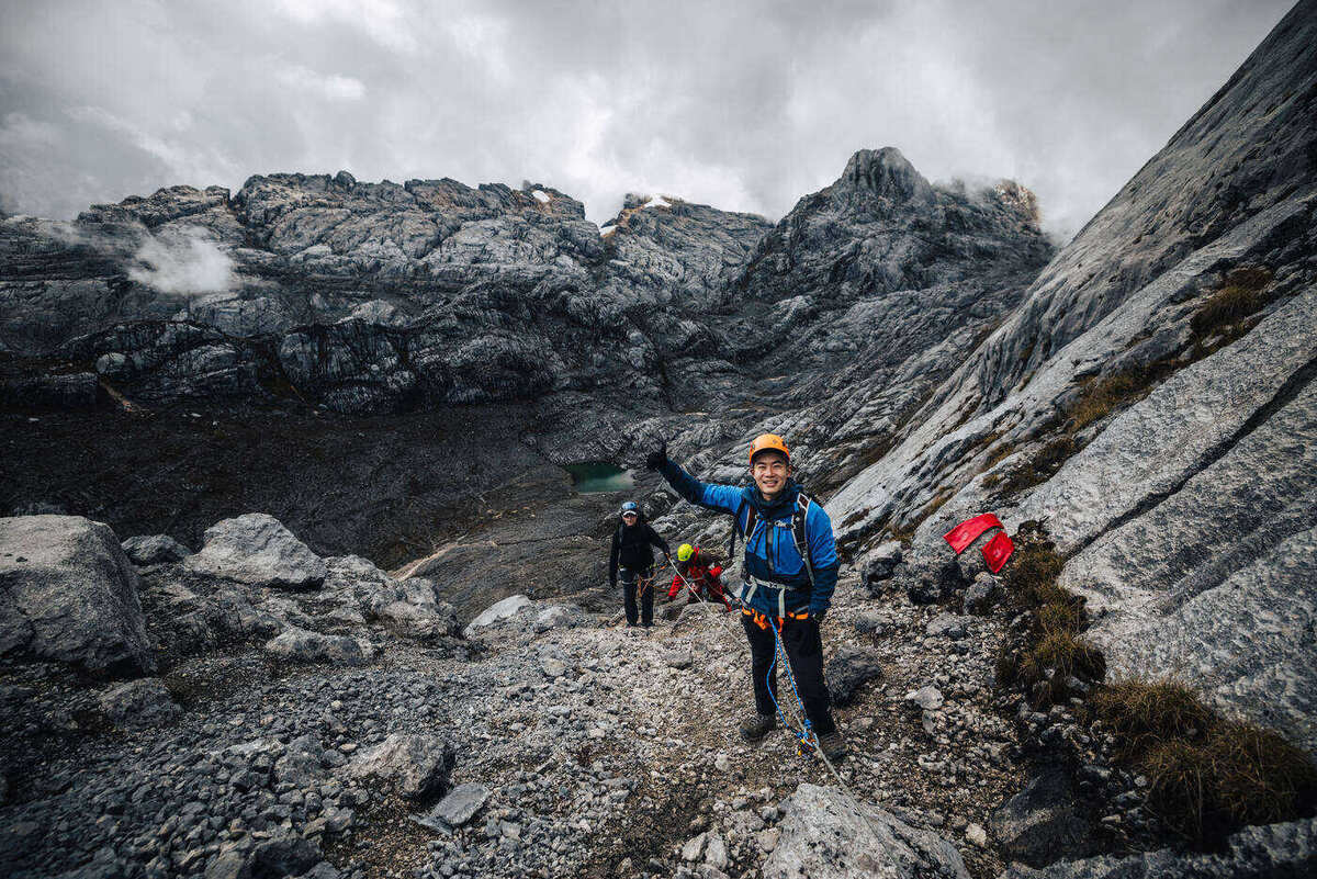Climber in blue jacket waving as the team walks up a rocky scree gully between limestone walls