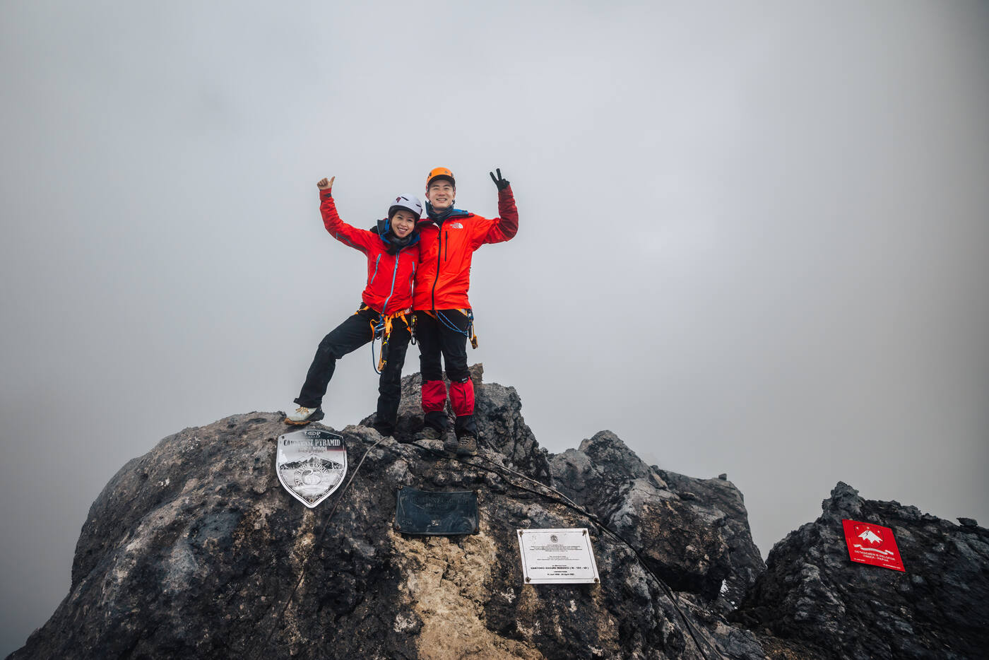 Two climbers in red jackets on summit with arms raised, misty mountains behind