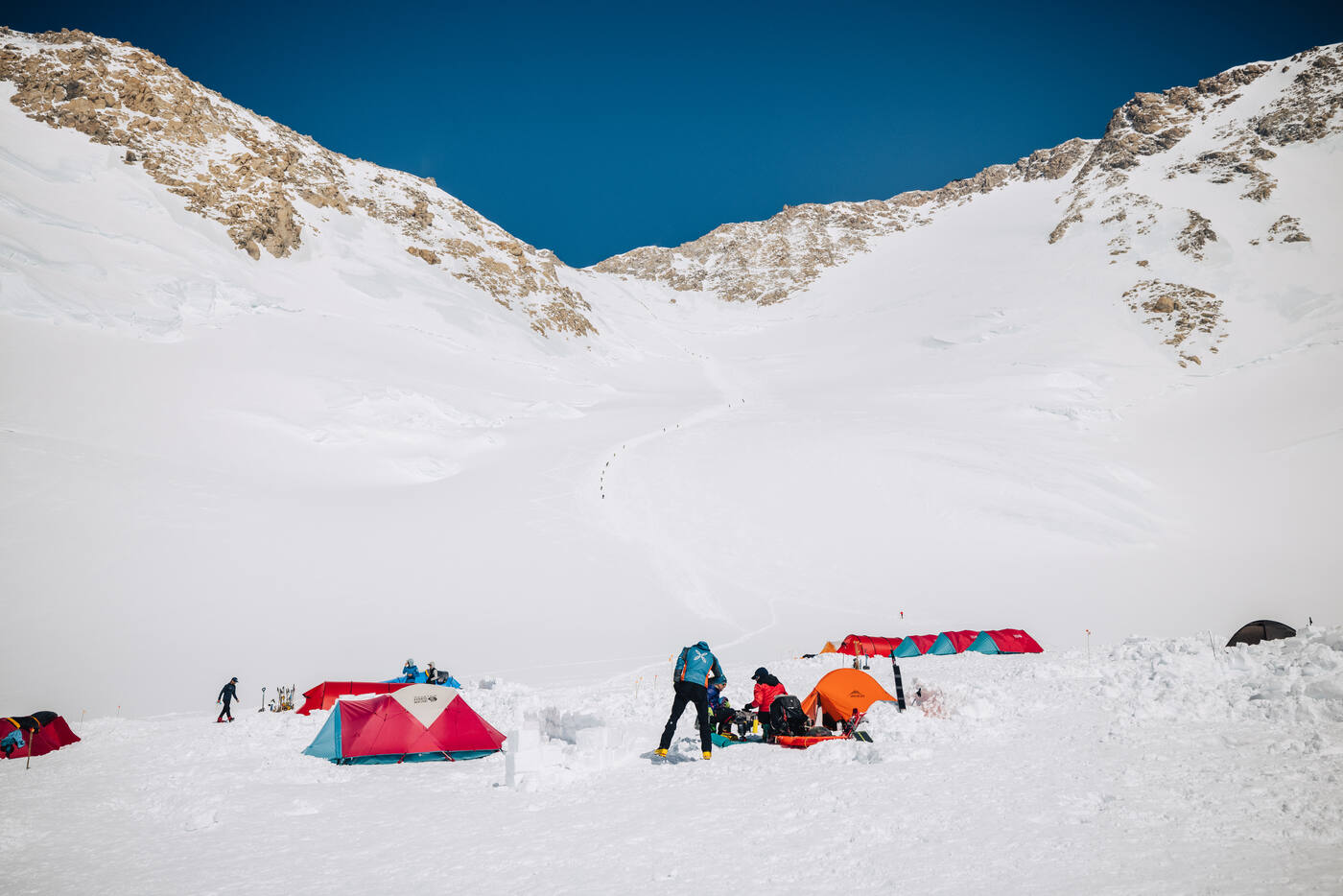 Tents pitched in the snow beneath a rocky ridgeline with a line of climbers ascending the slope above