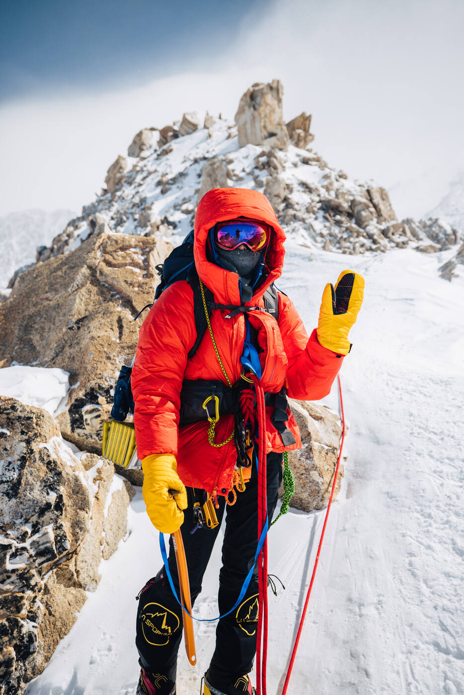 Climber in a red down parka and goggles waving from a rocky ridge with a rope trailing behind