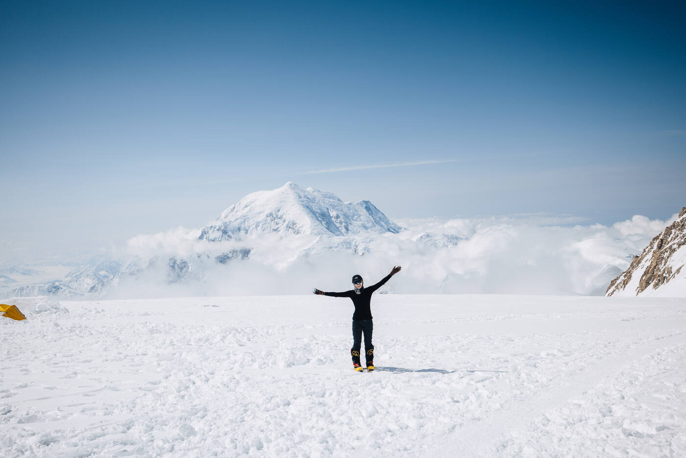Climber standing with arms spread wide at 14,200 ft camp with a massive snow-covered peak behind