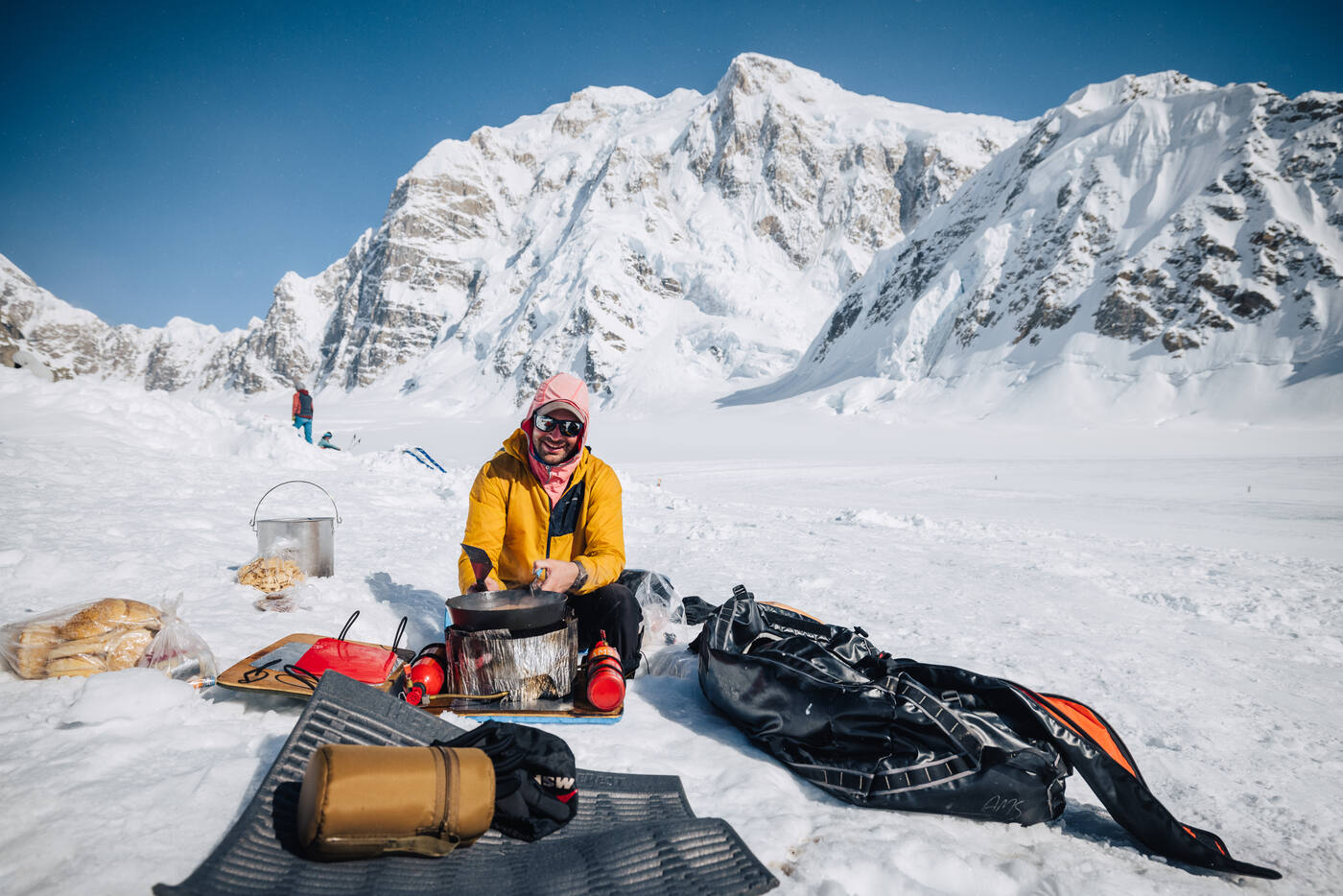 Climber in yellow jacket cooking at basecamp with Denali towering behind