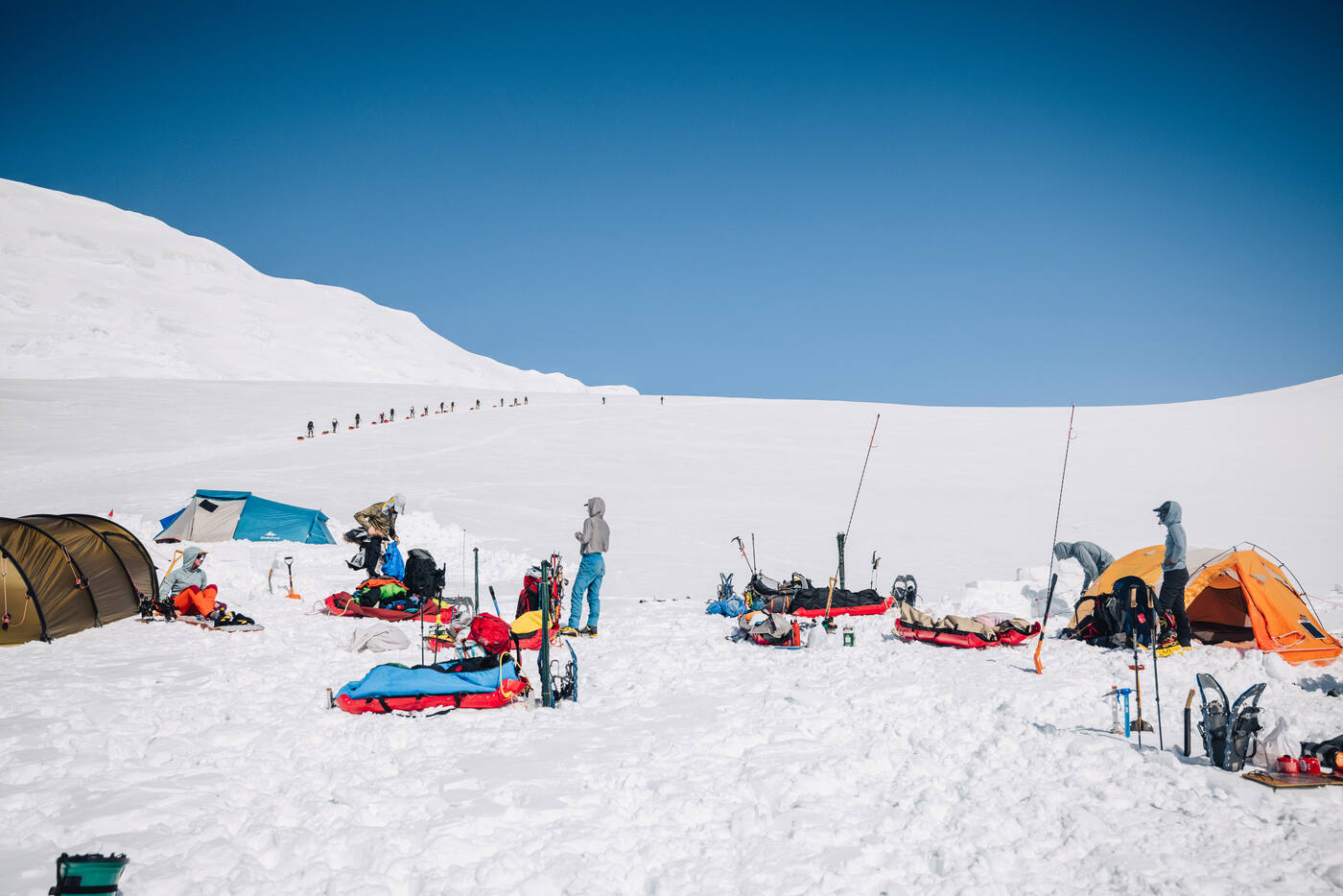 Tents and gear spread across a bright snow camp with a line of climbers ascending the slope behind