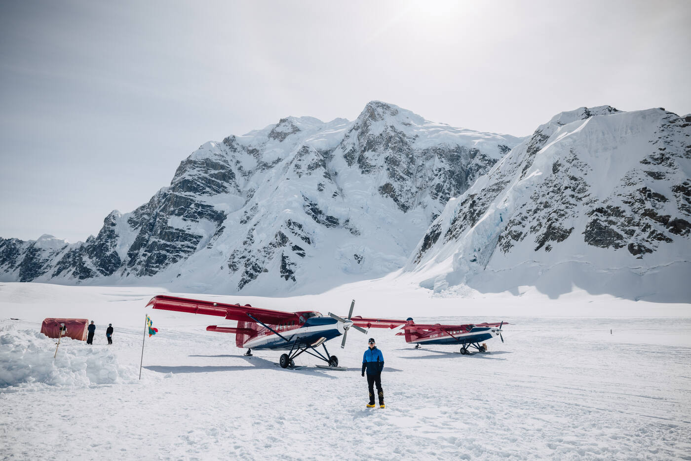 Two red bush planes parked on the Kahiltna Glacier landing strip with a climber standing nearby and snow peaks behind