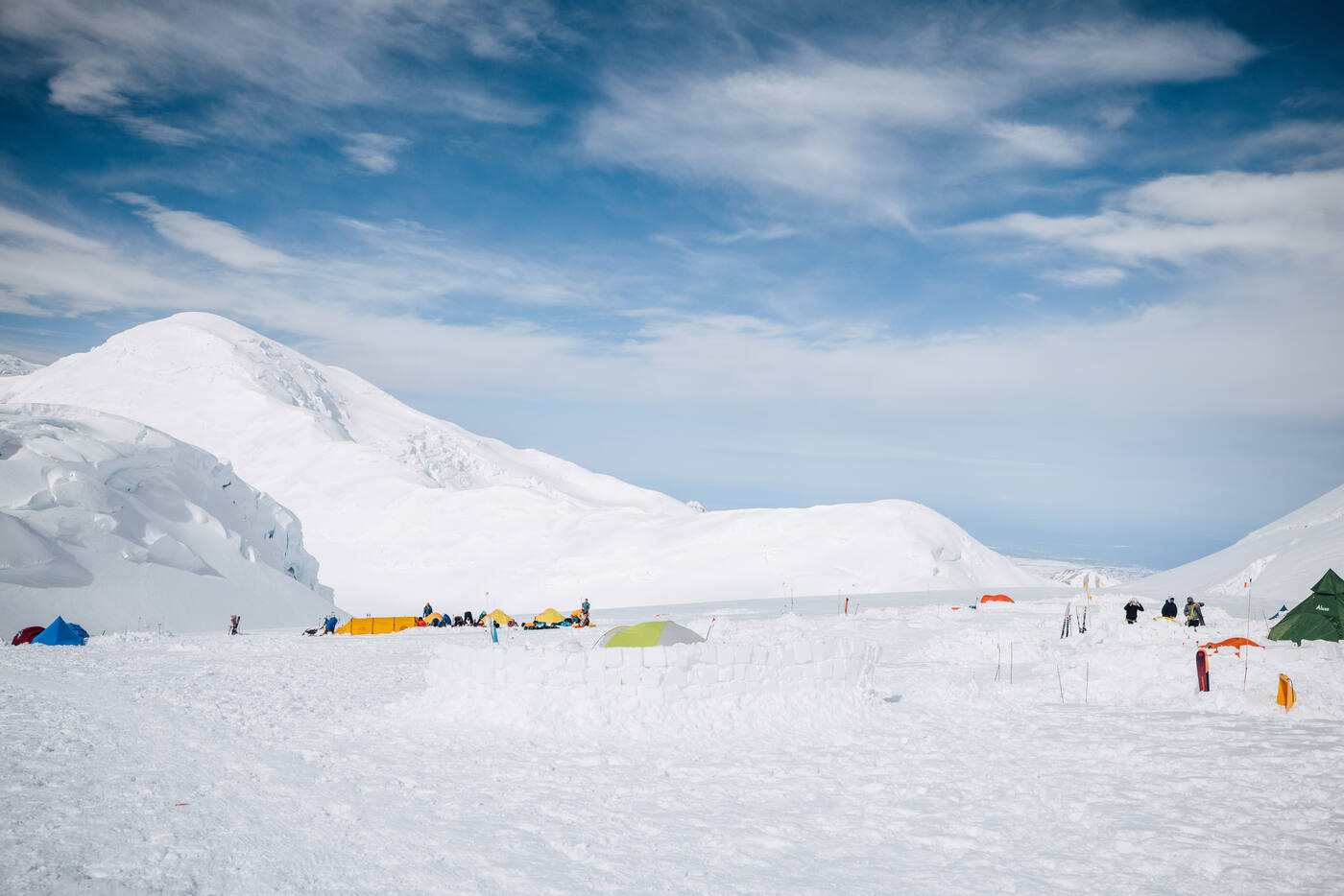 Tents scattered across the snow at camp beneath a broad snow dome and streaky clouds