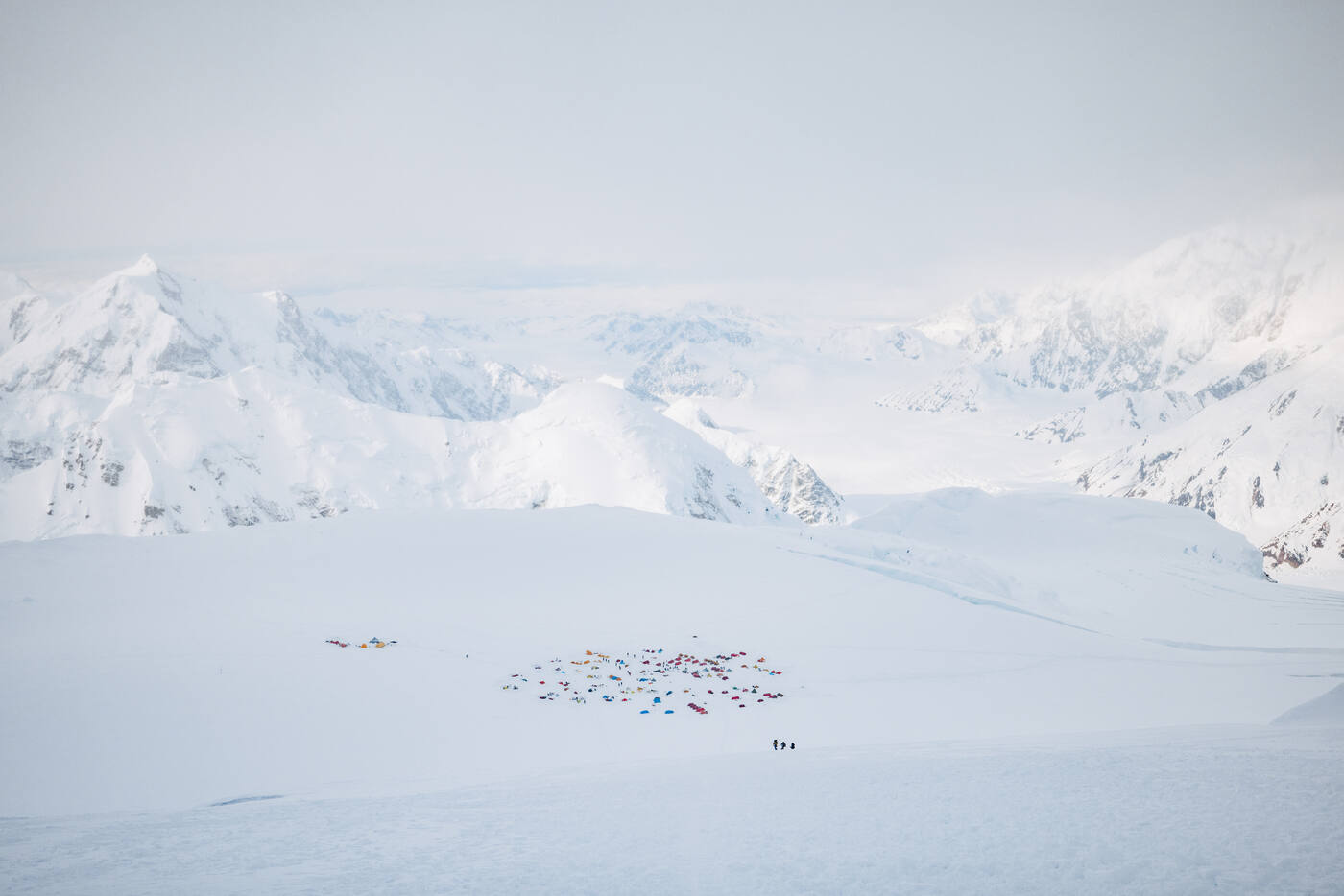 Distant view down onto a sprawling tent camp on the glacier with snow peaks and clouds beyond