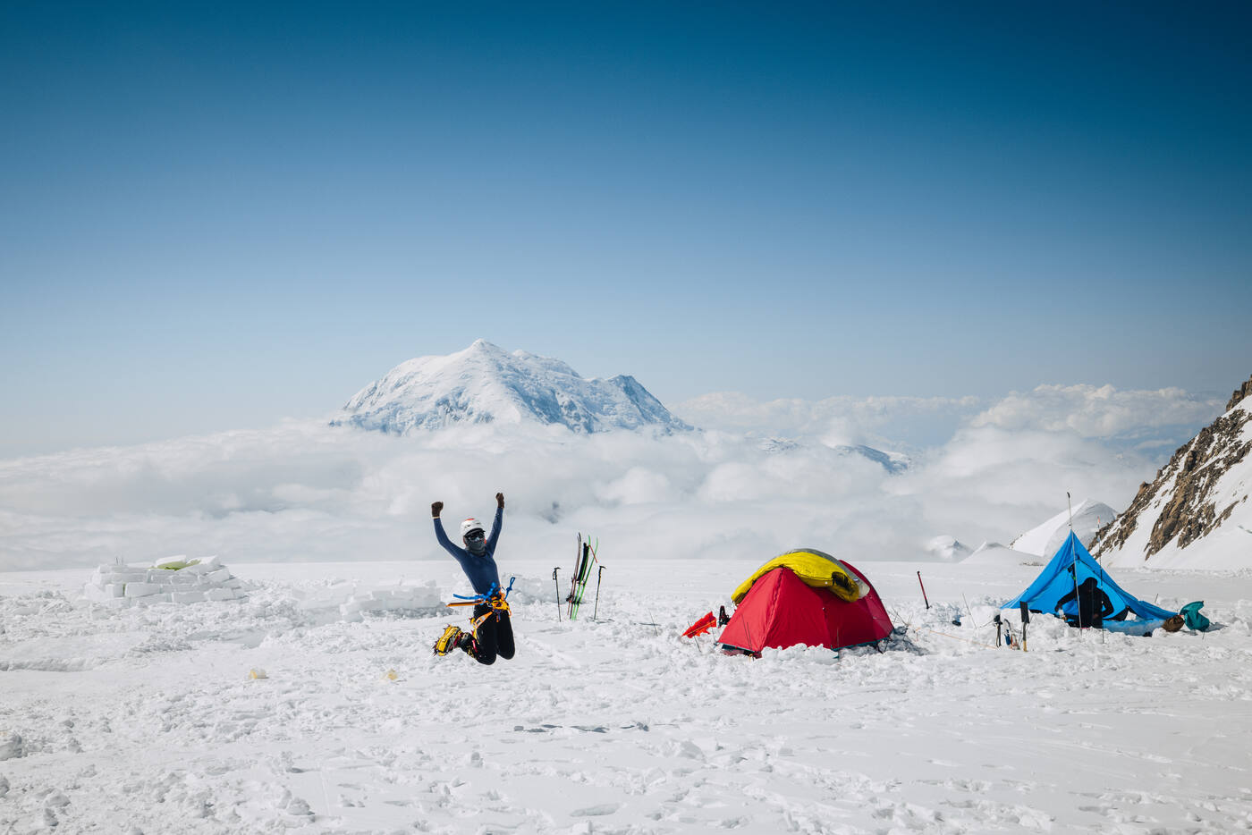 Climber jumping for joy at camp after summiting Denali with tent and mountains behind