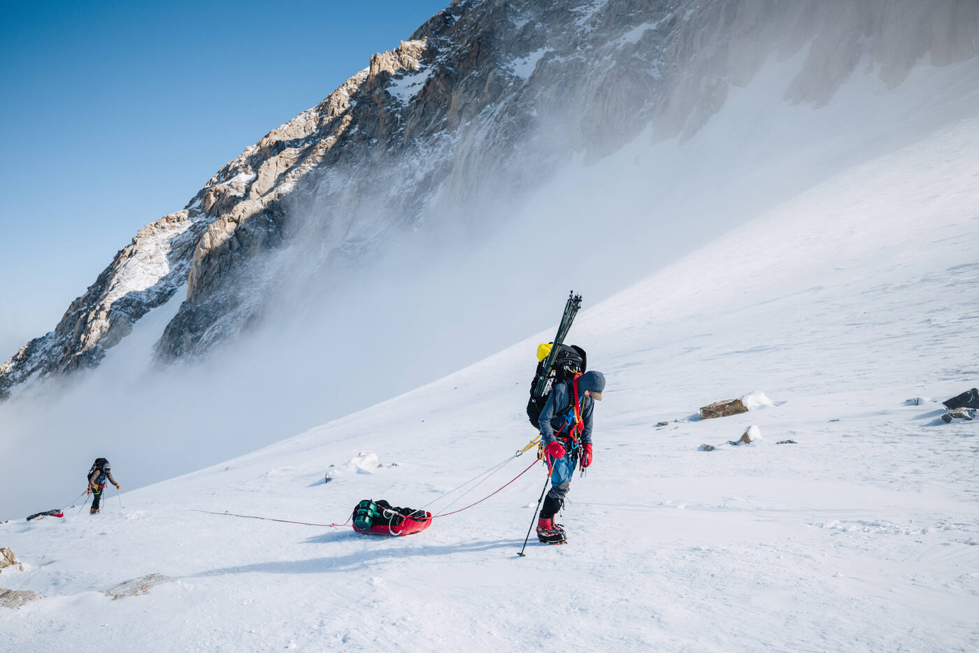 Climber hauling a loaded sled up a steep snow slope with rocky face above and wind-blown snow