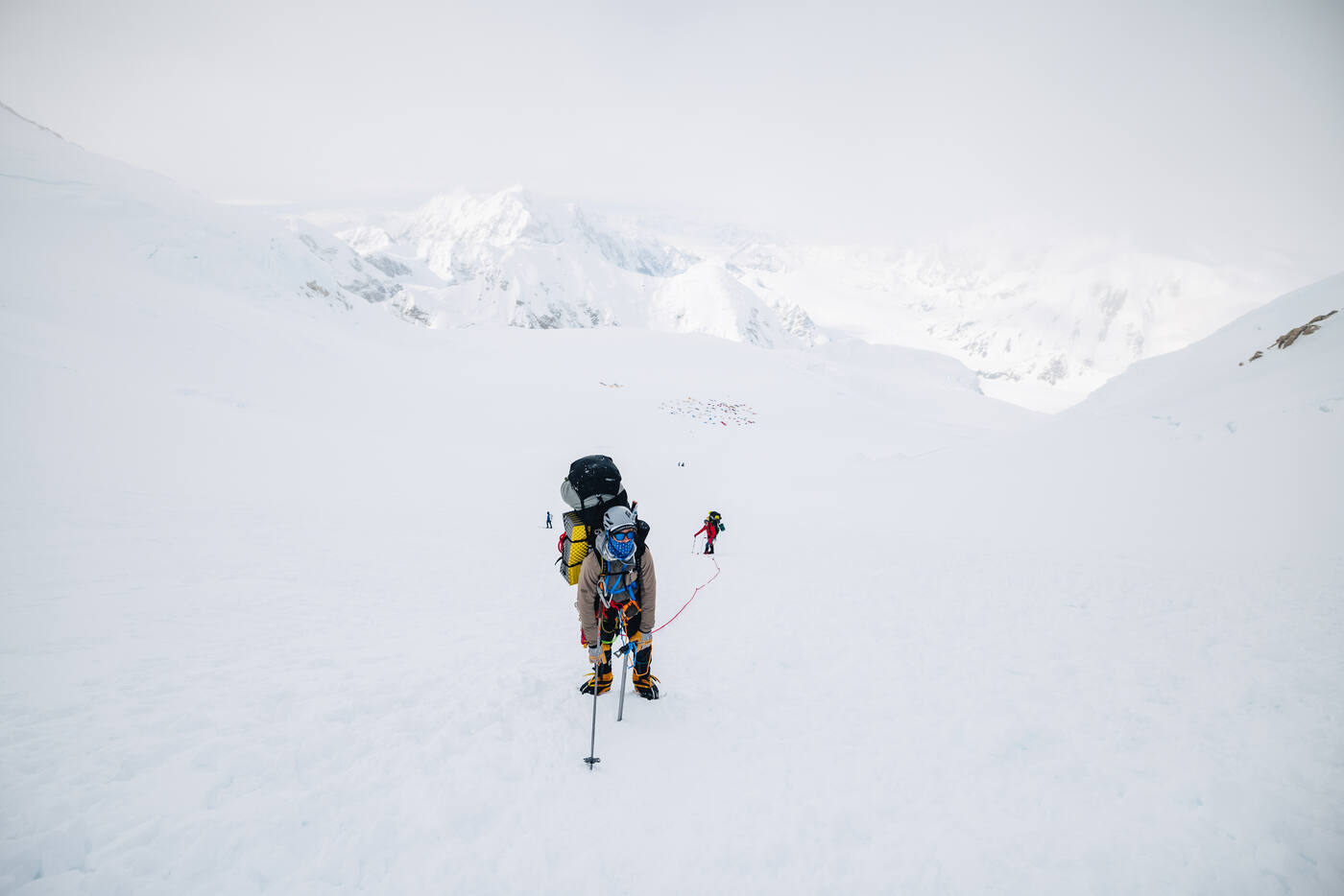 Climber with a heavy pack and trekking poles crossing a broad snow slope under flat overcast light, a distant camp below