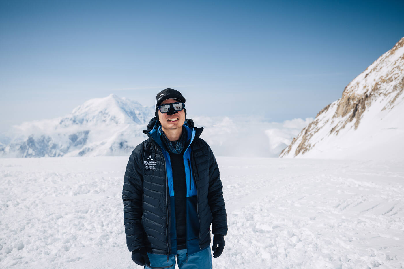 Climber in a black puffy jacket, cap, and sunglasses posing at camp with a snow peak and clouds behind