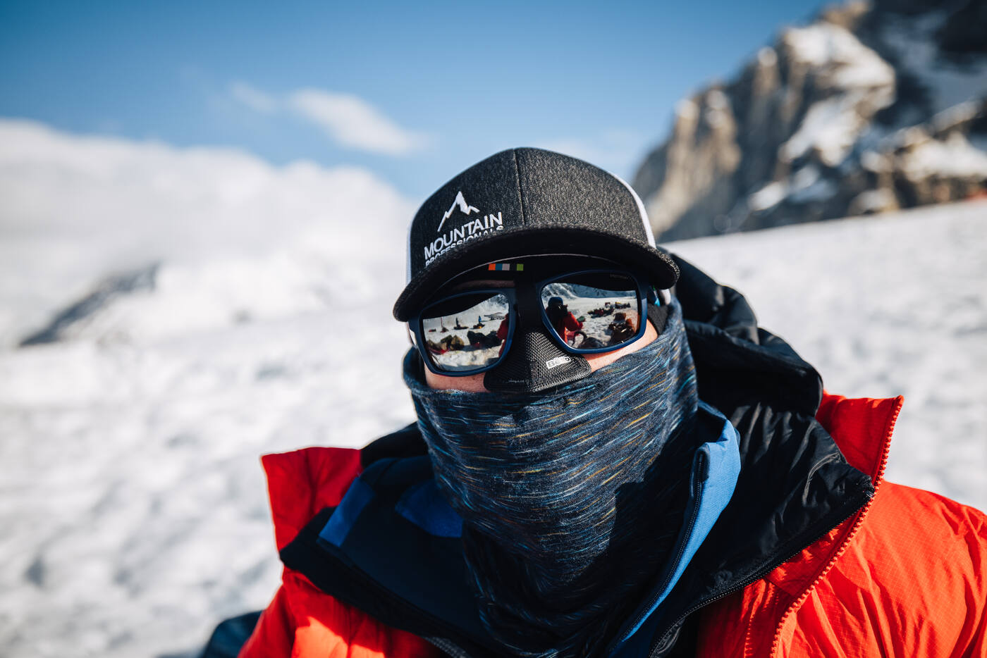 Close-up of a climber in a cap, mirrored sunglasses, and neck gaiter wearing a red parka with rocky peaks behind