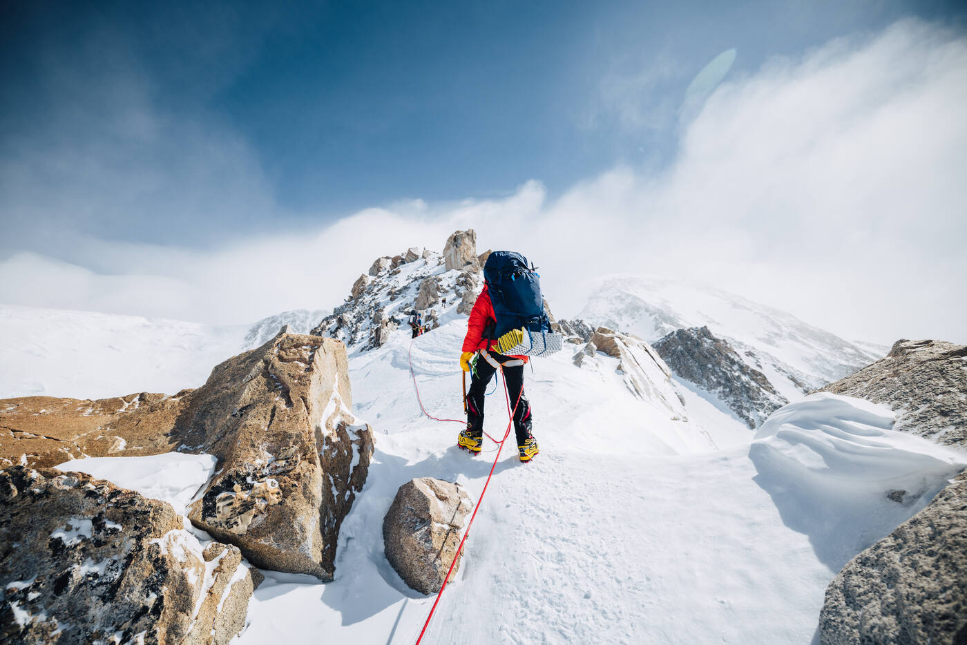Climber on a narrow rocky ridge with Denali's upper slopes visible through clouds