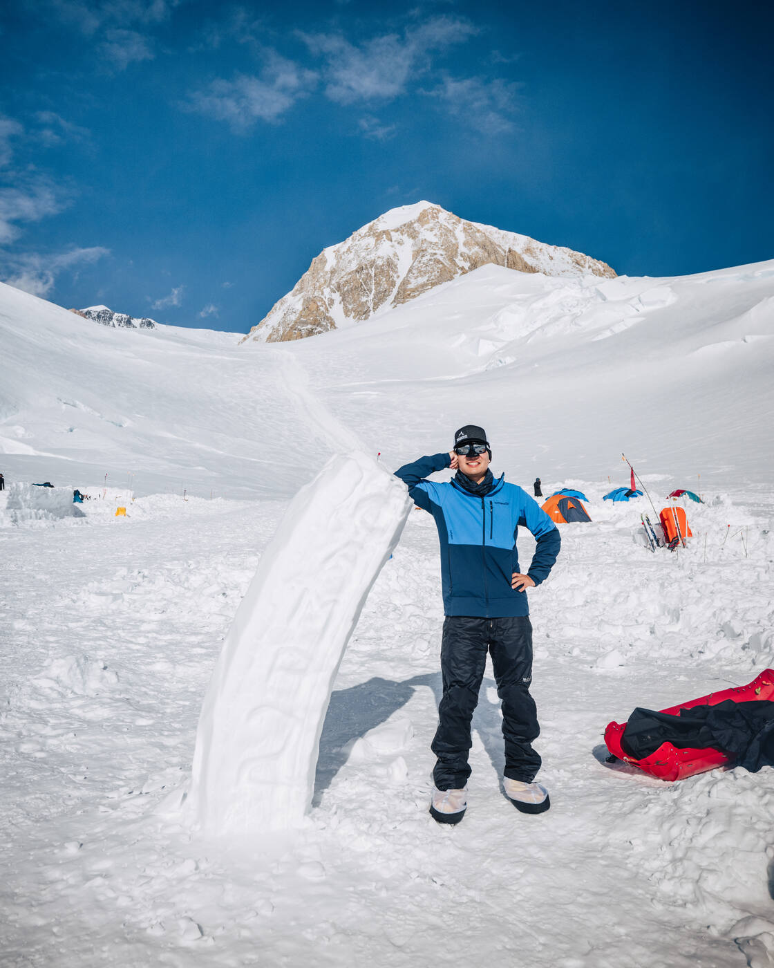 Climber in a blue jacket saluting beside a carved snow block at camp with a rocky peak behind
