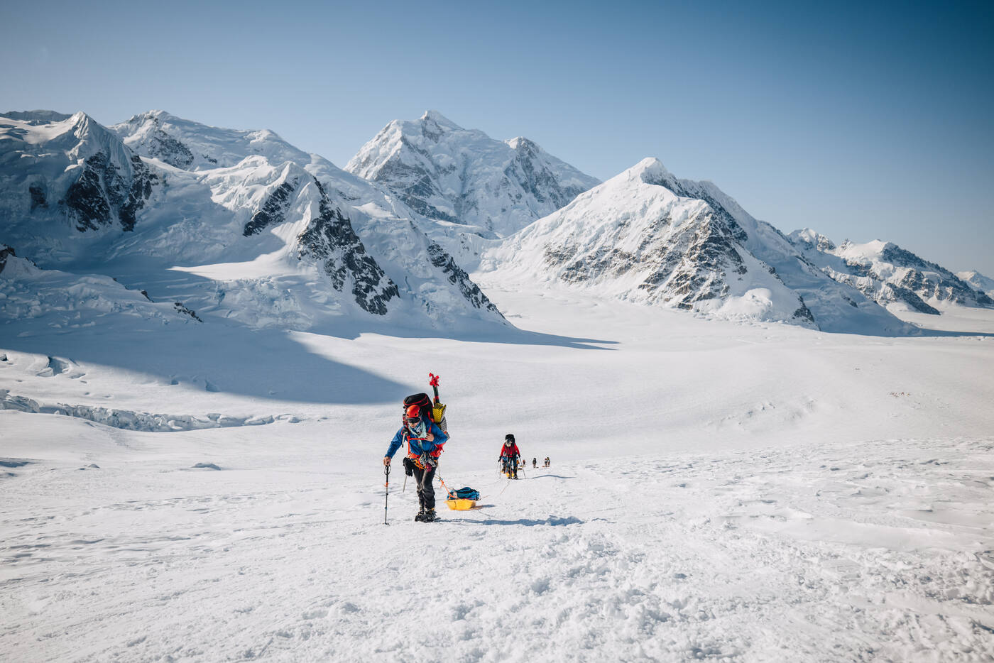 Climber with a heavy pack and sled crossing a gentle glacier with jagged snow-covered peaks behind