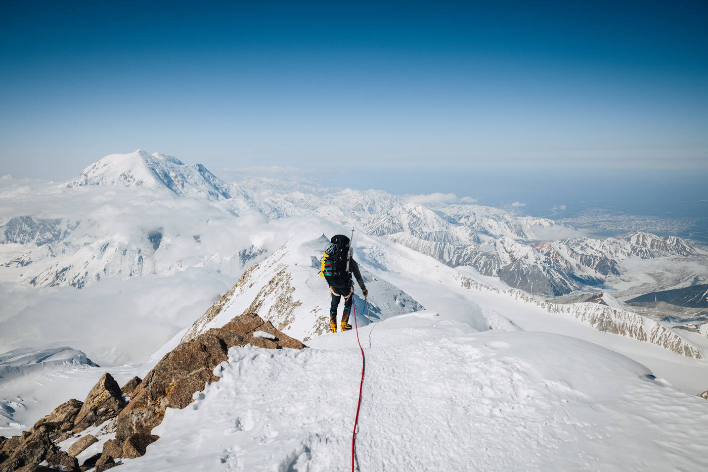 Climber on a rocky ridge with a rope trailing behind, snow peaks and clouds stretching beyond