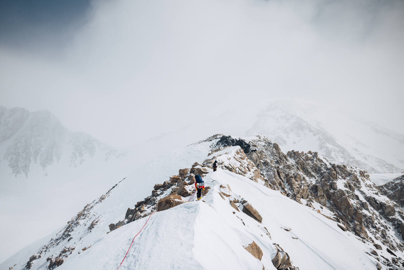 Climbers traversing an exposed snowy ridge high on Denali with mountains and clouds behind