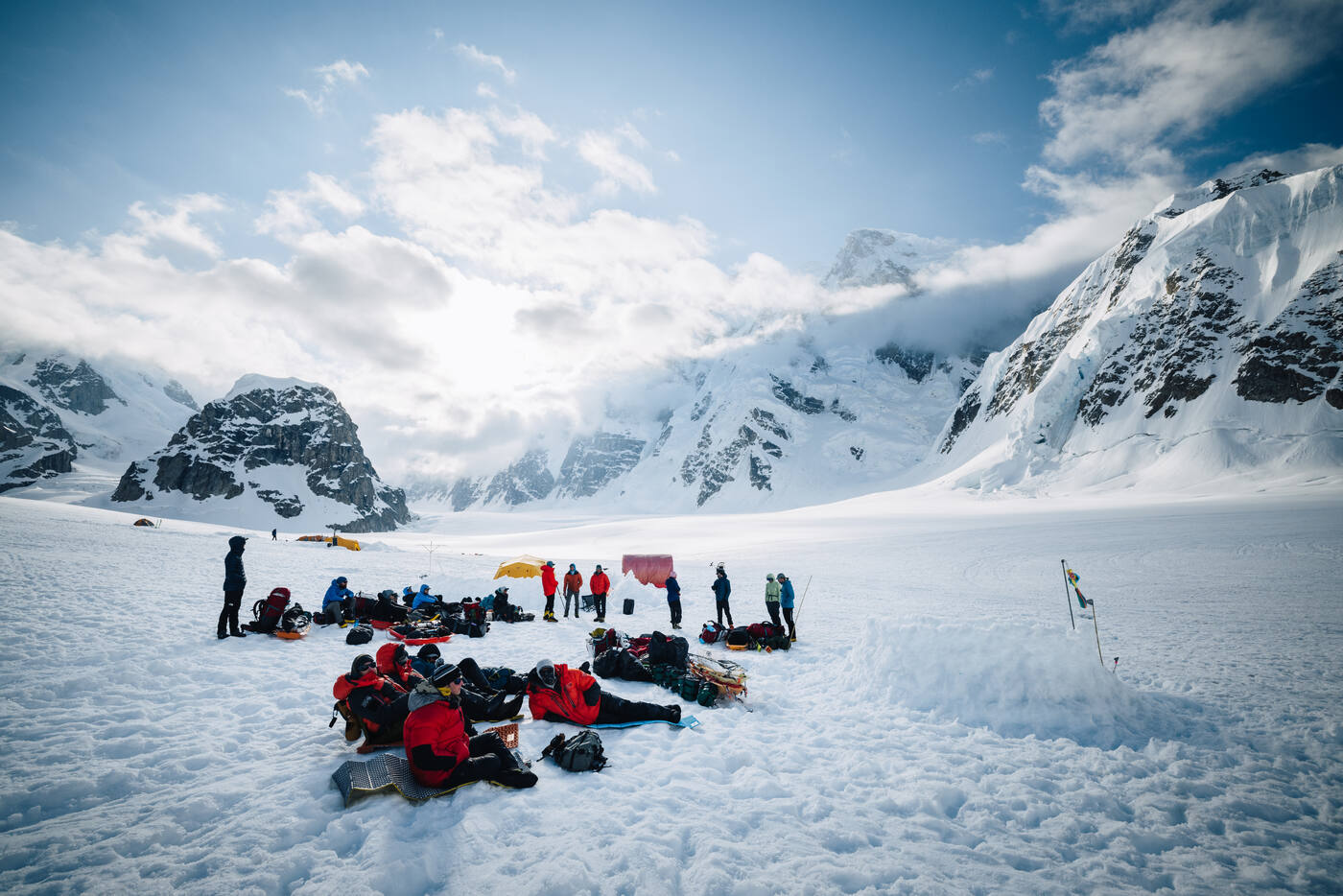 Climbers sorting gear and packing up at camp in the morning with sunlit peaks and clouds behind