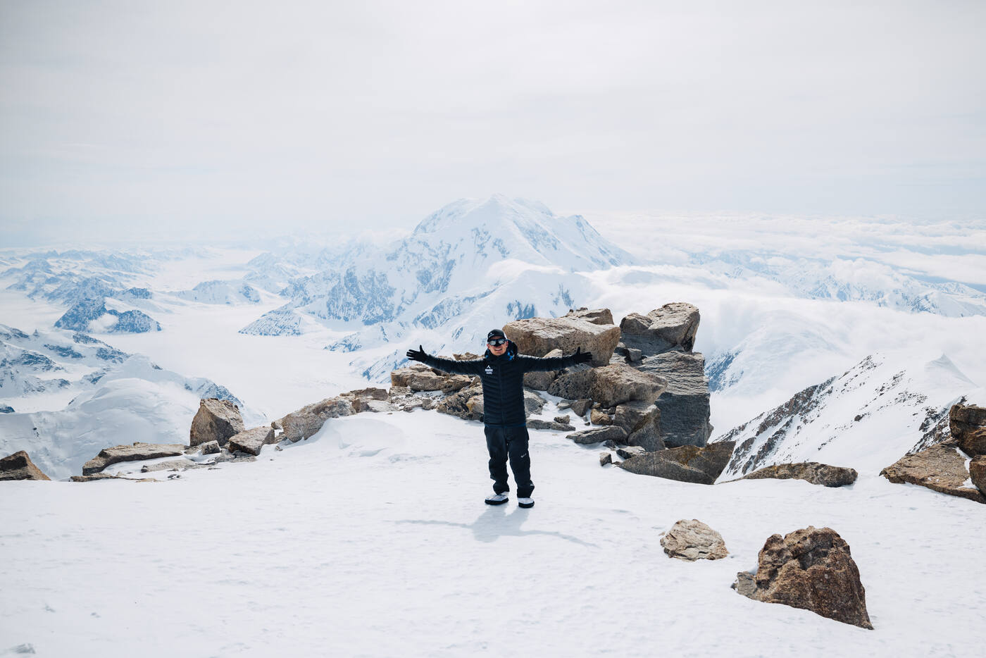 Climber standing with arms outstretched on rocky outcrop at high camp with Alaska Range panorama