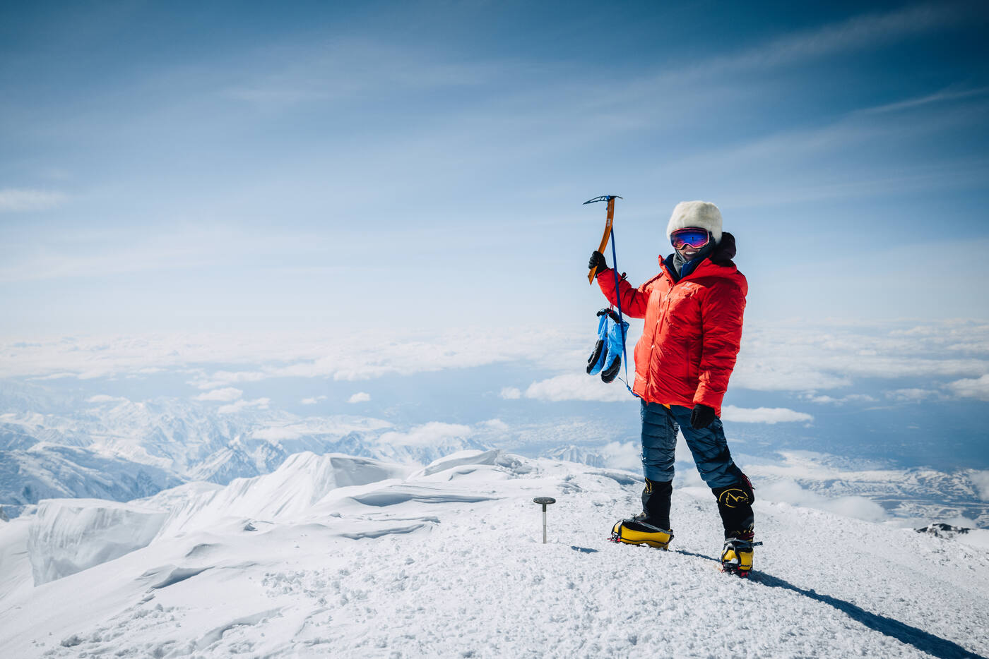 Grace standing at the summit raising ice axe overhead with mountains below