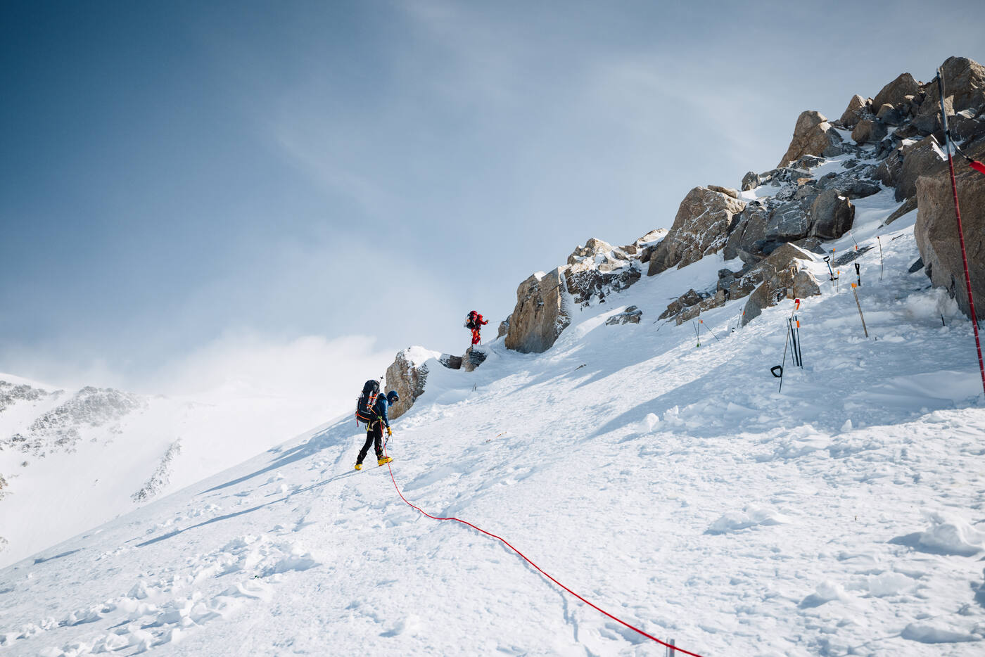 Climbers clipped to fixed lines on a steep snow slope beneath a rocky outcrop, with anchors and wands marking the route