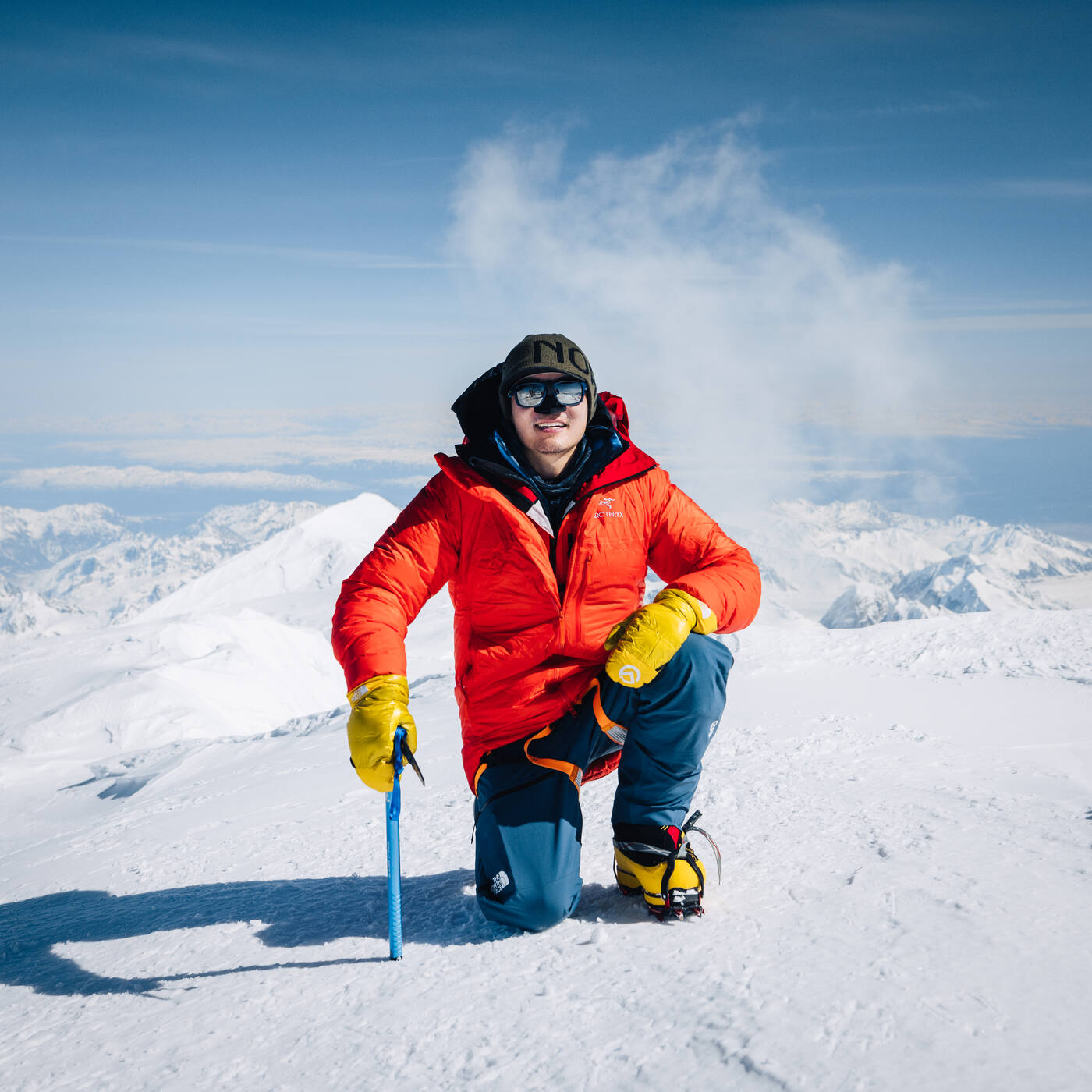Jason kneeling at the summit of Denali in red jacket with ice axe, Alaska Range panorama behind