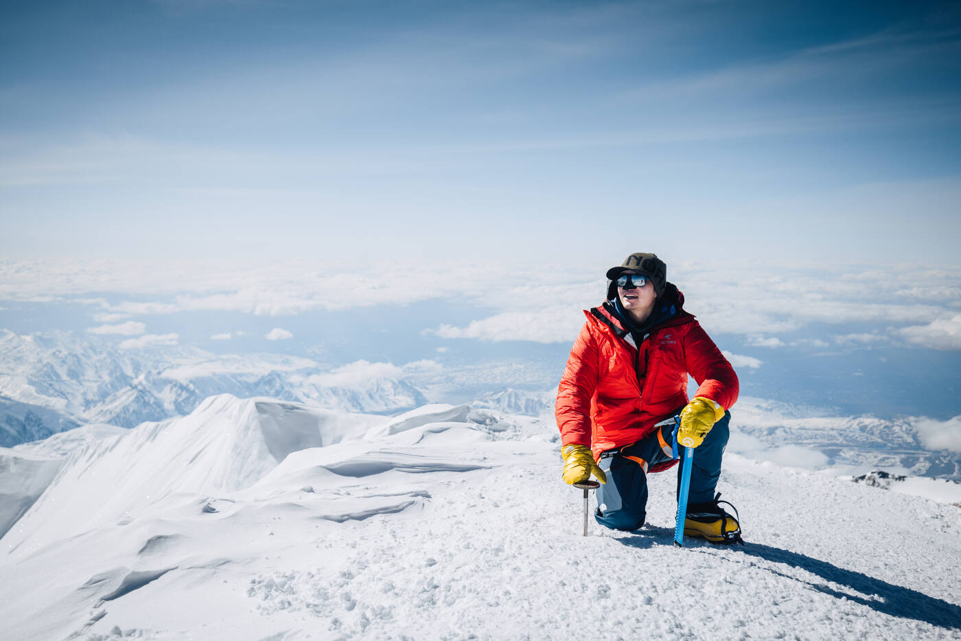 Jason kneeling at the summit with ice axe and sunglasses, mountain panorama behind