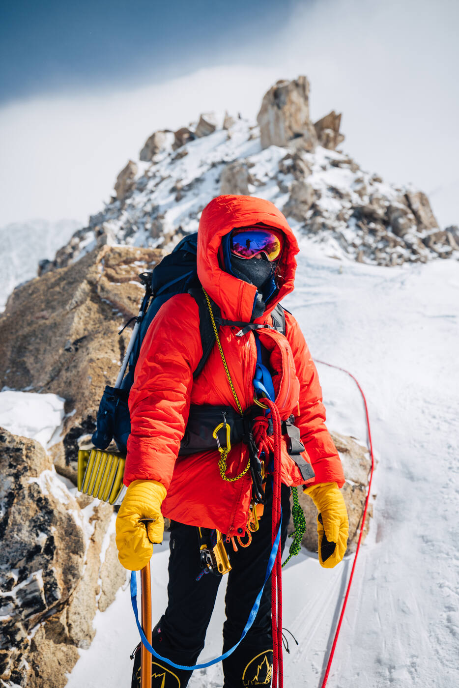 Climber in a red down parka and goggles standing on a rocky ridge with a rope clipped in