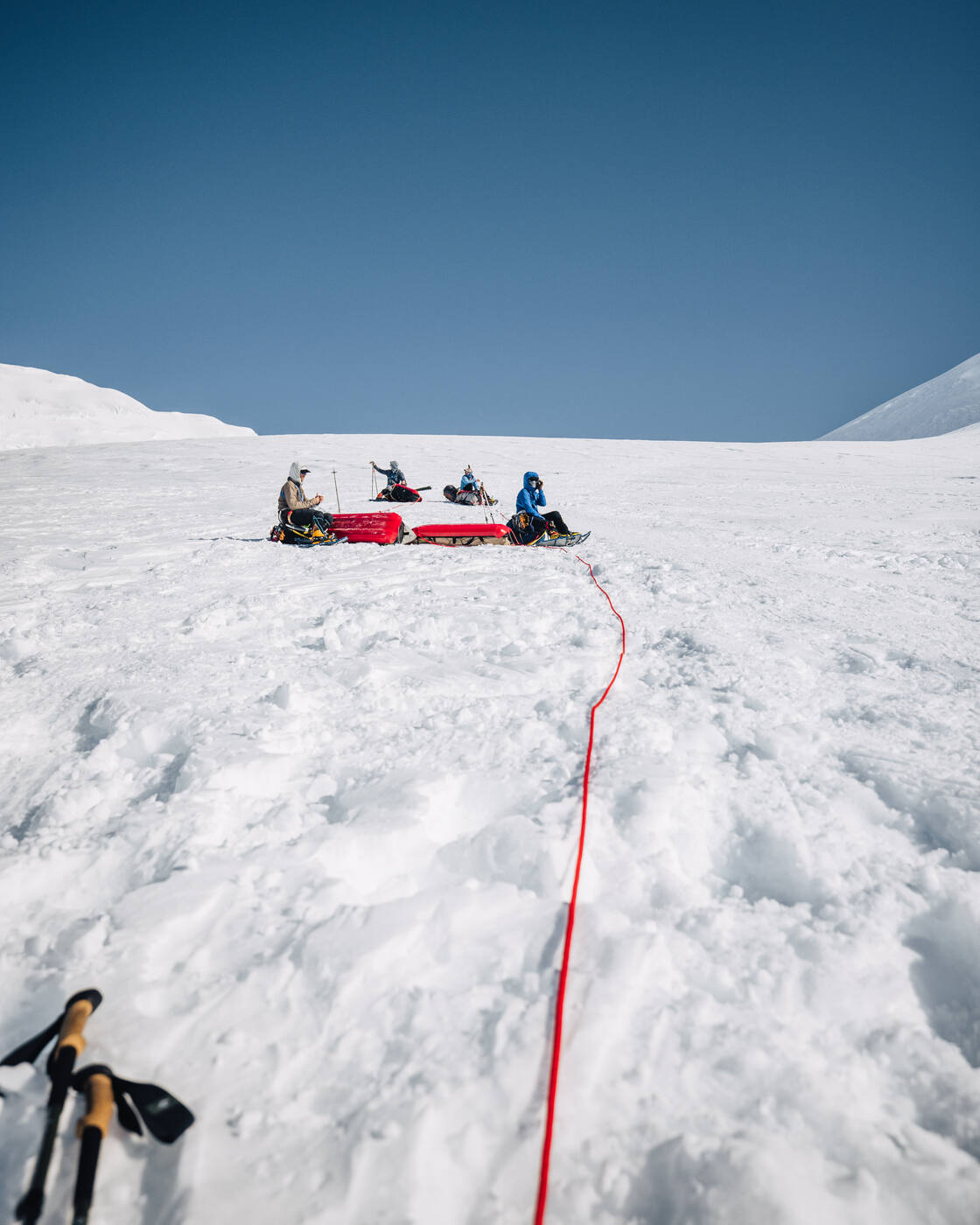 Rope team taking a break with their sleds on the glacier, red rope trailing back to trekking poles in the foreground