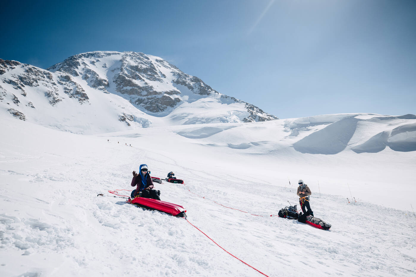 Climbers resting beside their sleds on a snow slope beneath a rocky peak