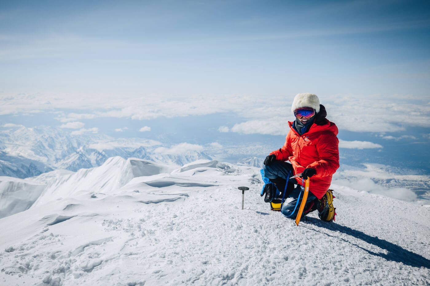 Climber in a red parka kneeling at the summit with the Alaska Range and clouds spread out below
