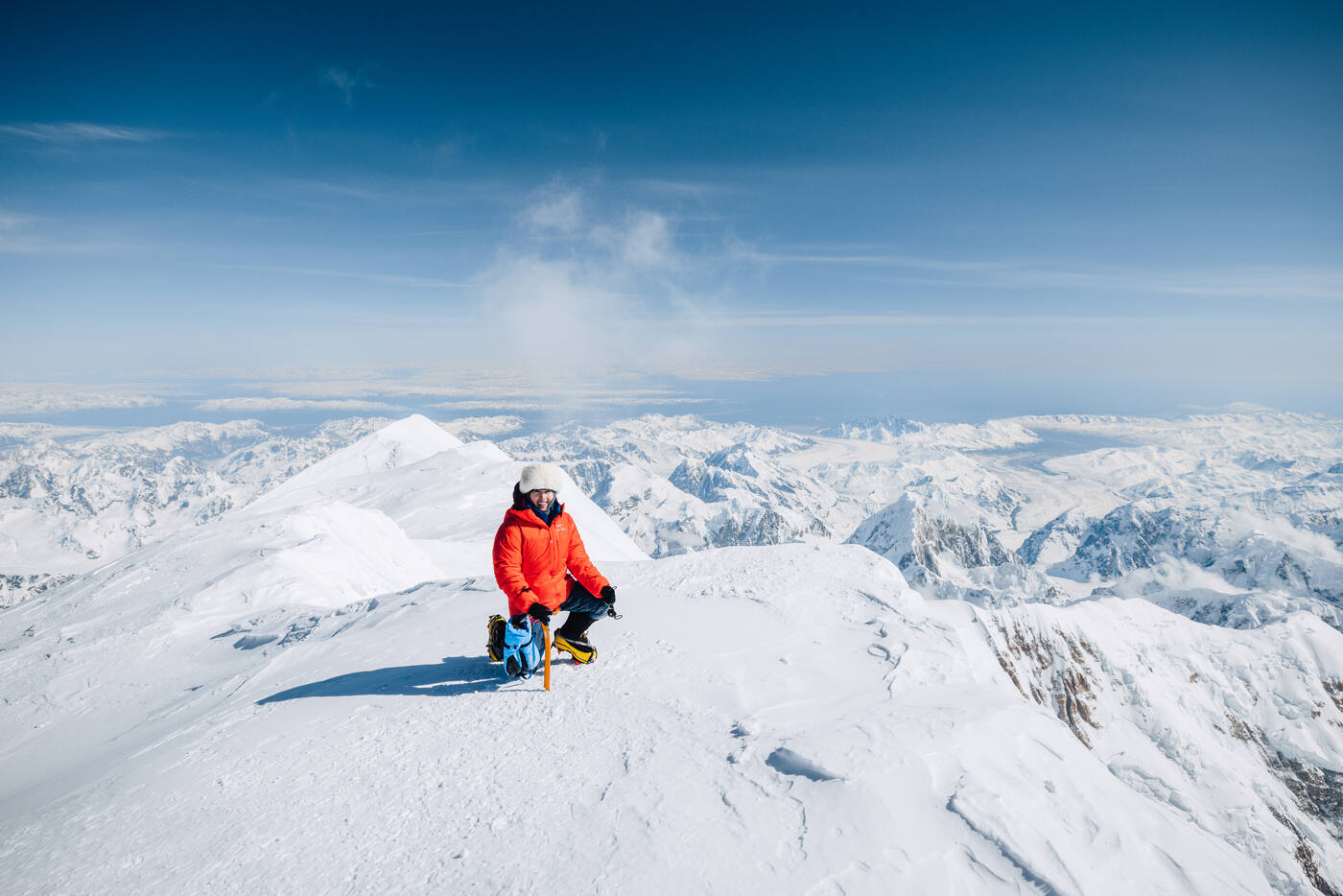 Climber in a red parka kneeling on the summit with peaks and clouds stretching to the horizon