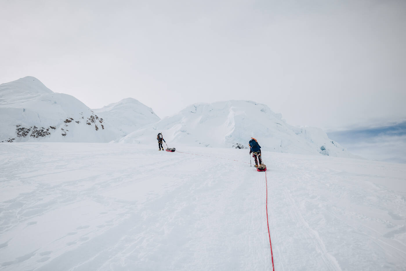 Two roped climbers hauling sleds across a flat section of glacier in flat grey light