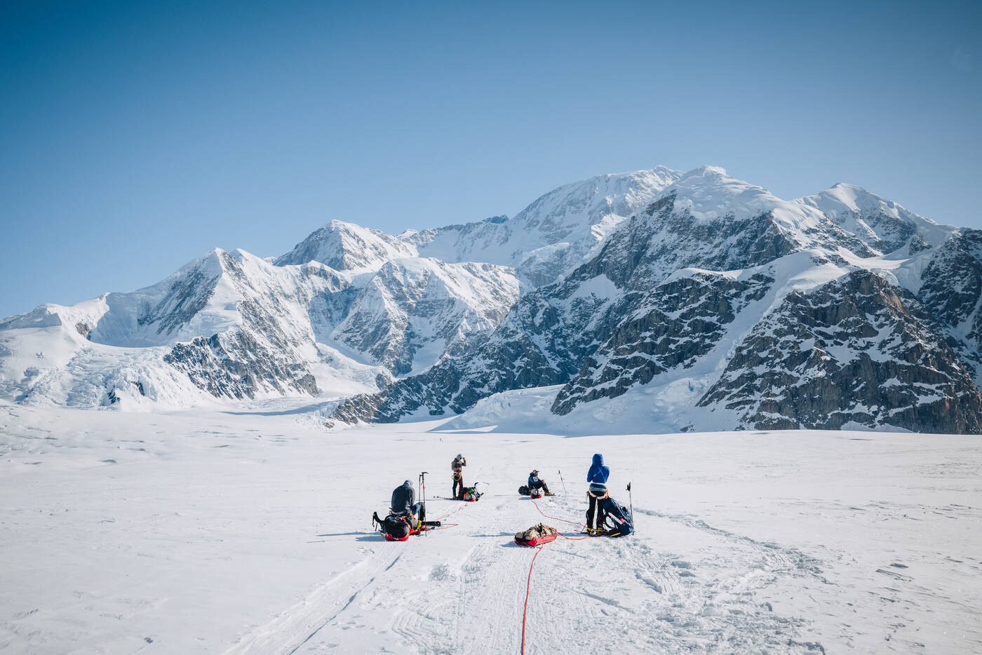Rope team resting beside their sleds on the flat glacier beneath towering snow-covered peaks