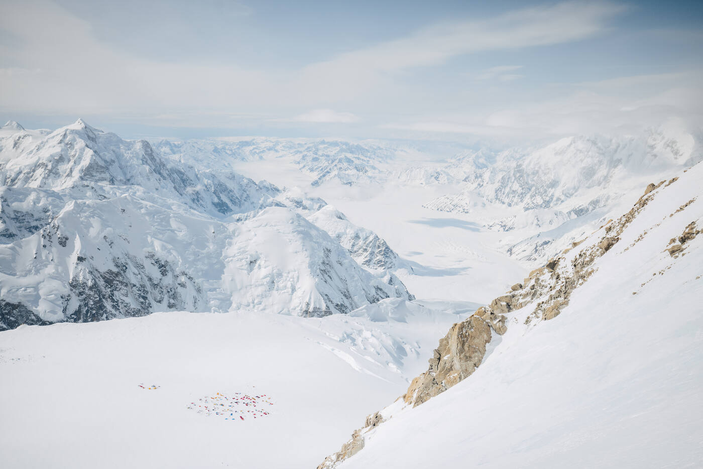 View down from the route to a cluster of colorful tents far below, with snow peaks and glaciers stretching to the horizon
