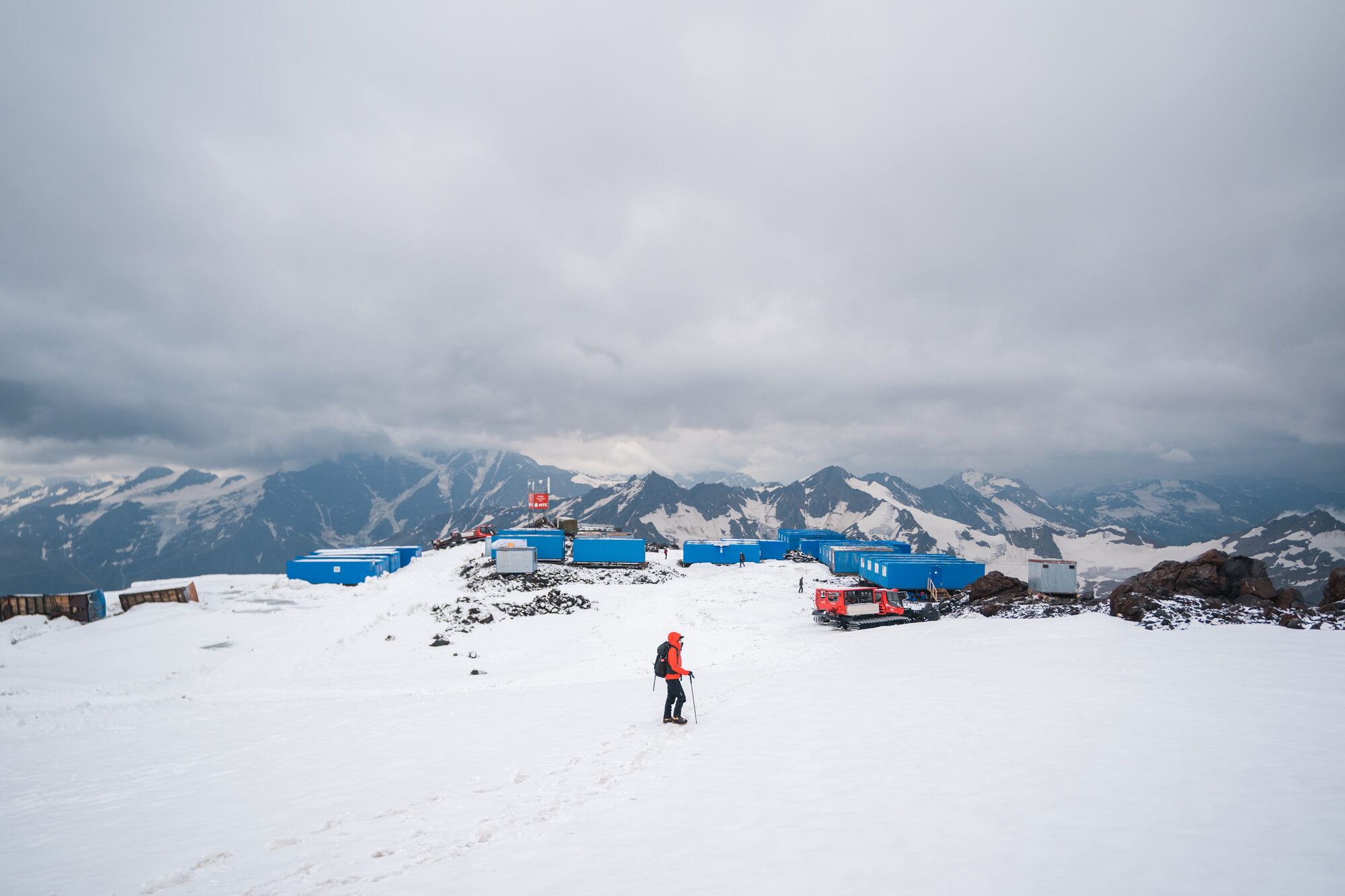 Lone climber walking across the snow toward the blue container huts of base camp with the Caucasus behind