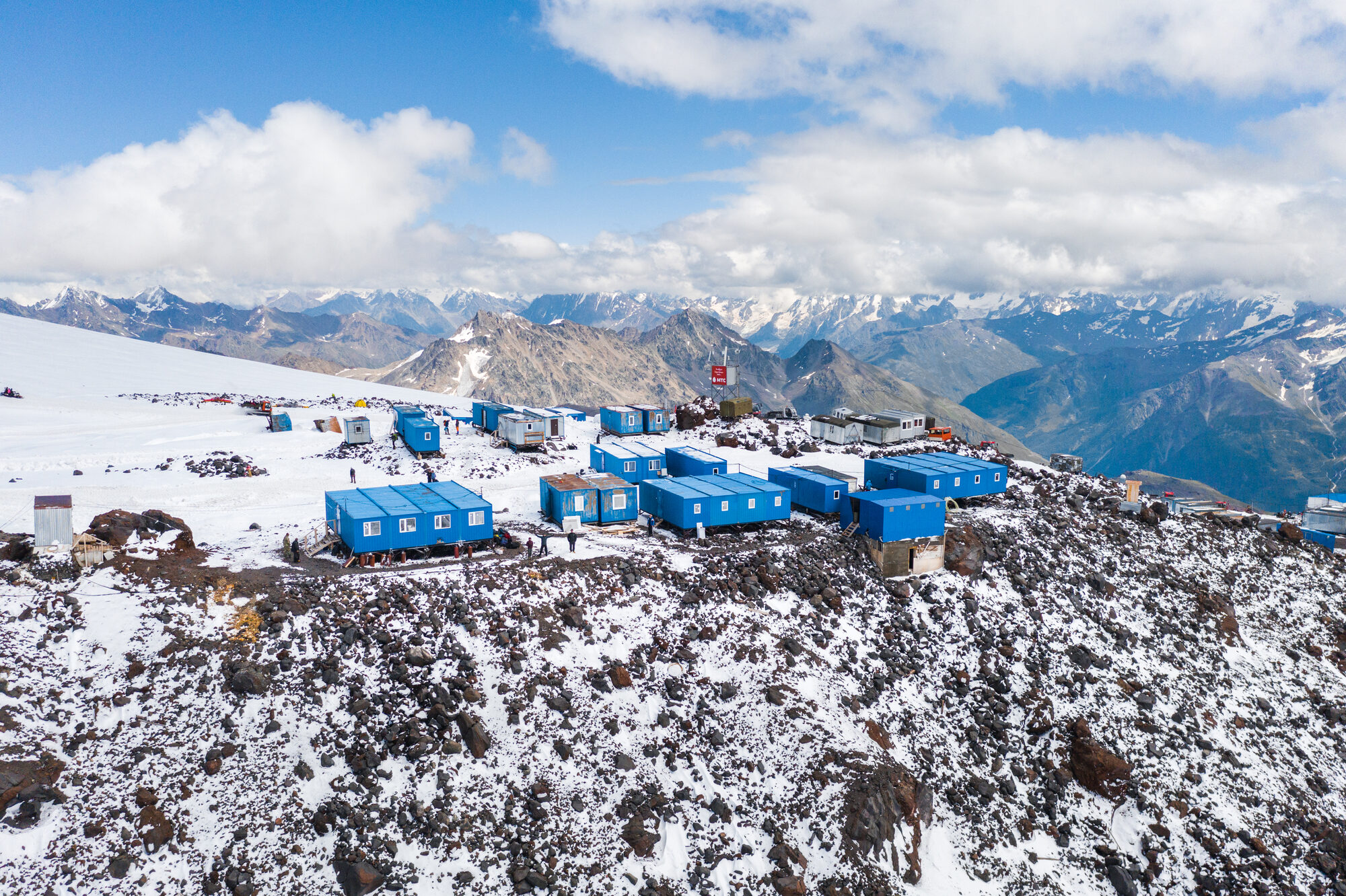 Drone view of the blue container huts at base camp on a rocky snow ridge with the Caucasus range beyond
