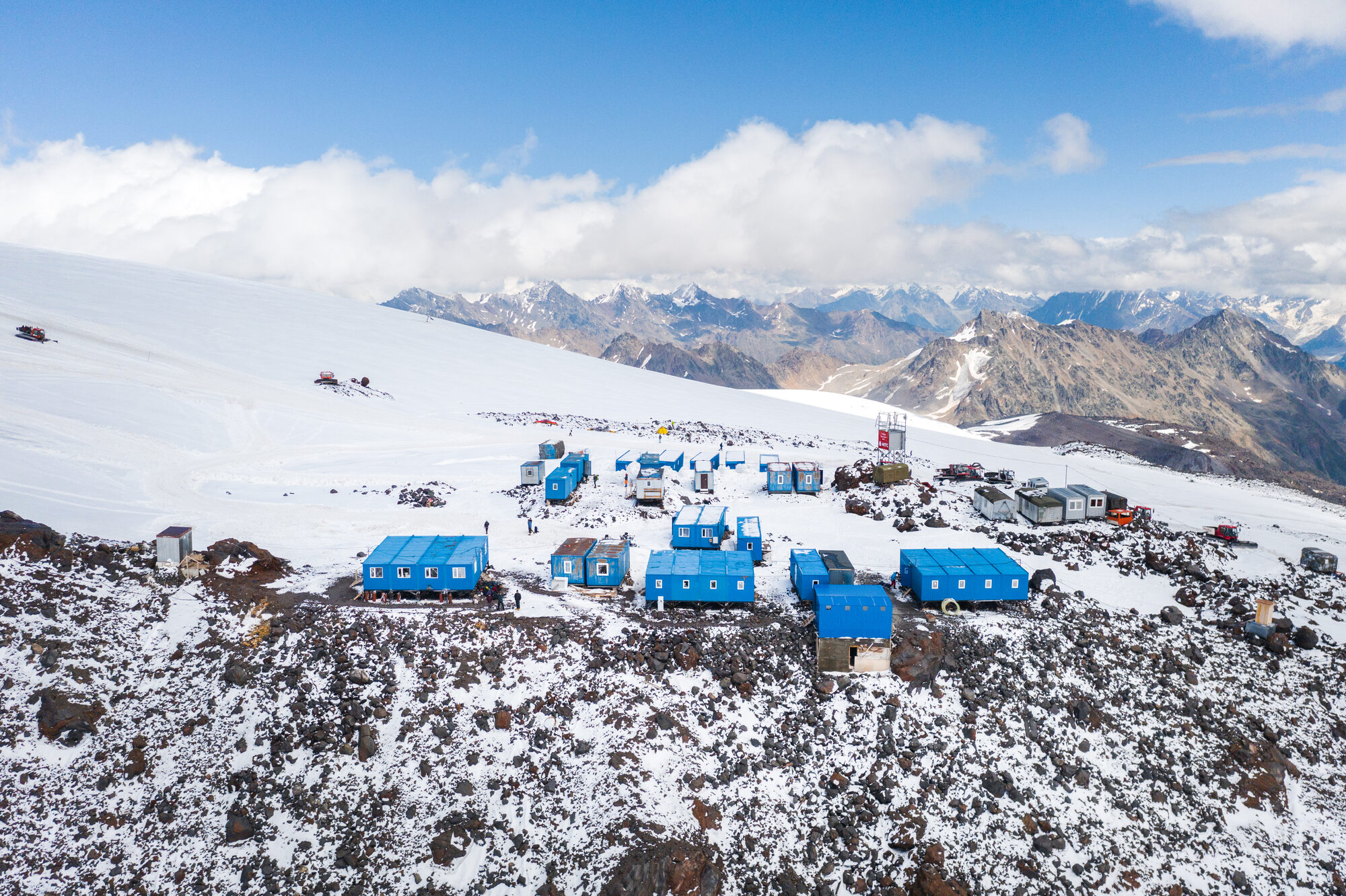 Wide drone view of the blue container huts and snowcats at base camp with the Caucasus range behind
