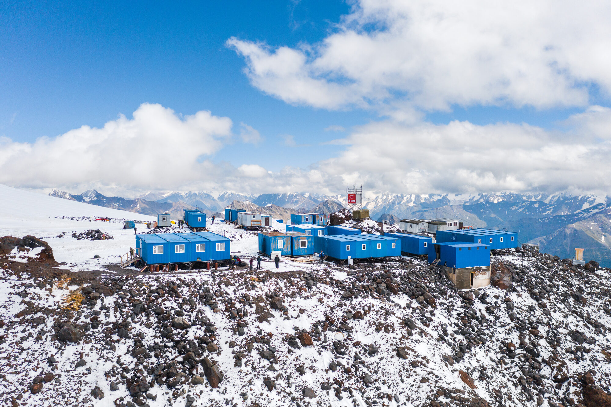 Wide drone aerial of the blue container base camp perched on a rocky snow ridge with Caucasus peaks behind