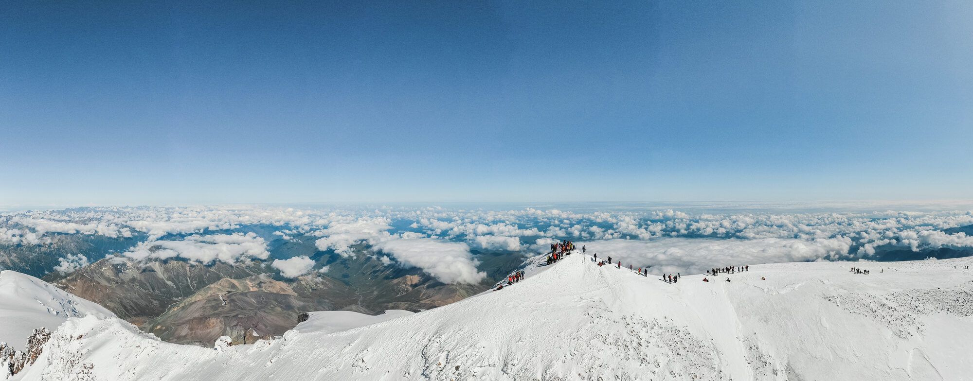 Drone panorama of a line of climbers along the snowy summit ridge of Elbrus above a sea of cloud