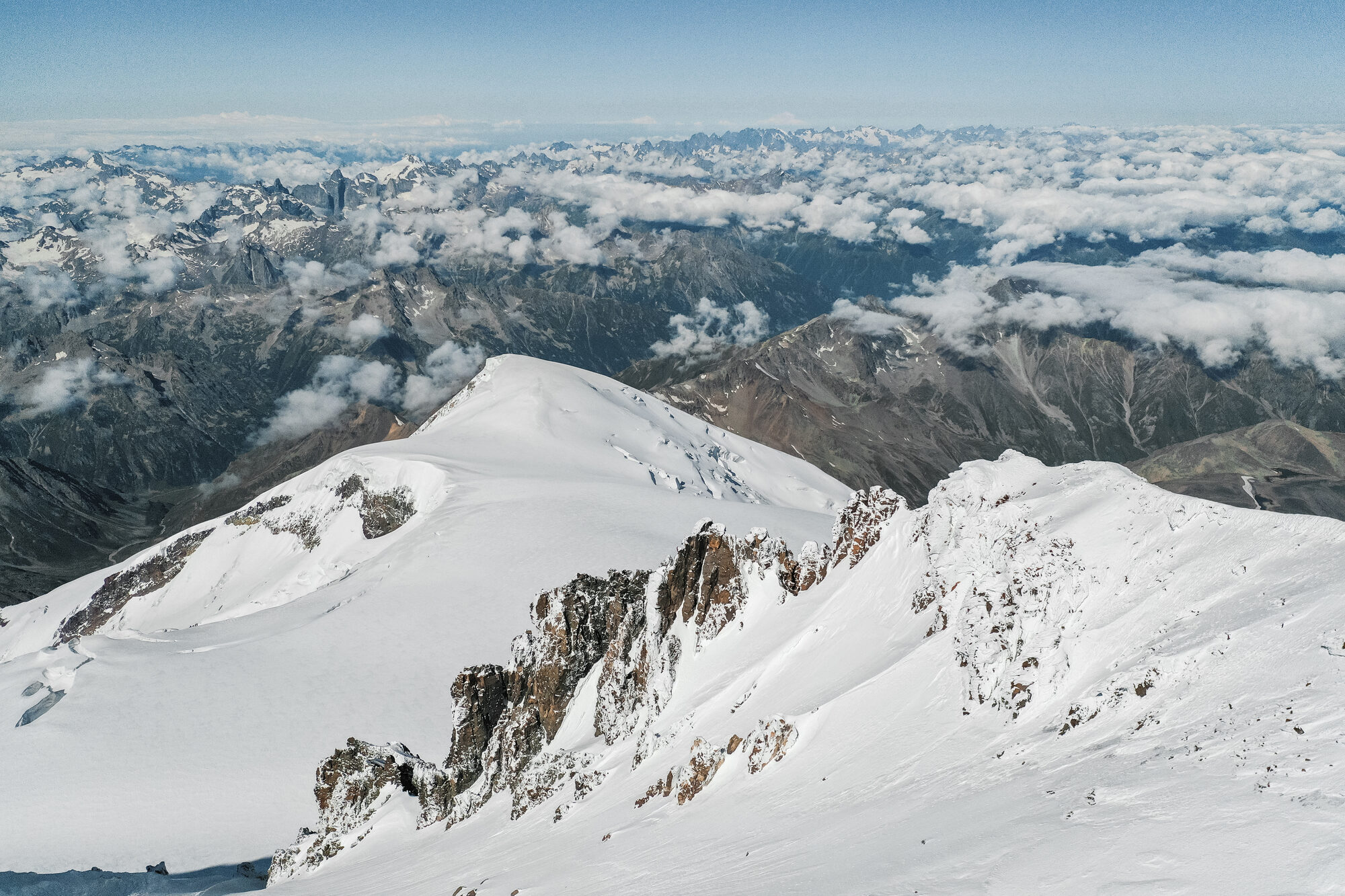 Drone view over the snow-covered summit dome and rocky ridge of Elbrus with the Caucasus and cloud beyond