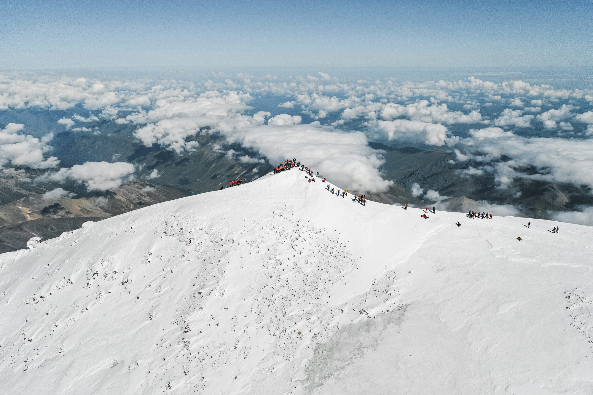 Drone aerial of the line of climbers descending the Elbrus summit ridge with shadow of the peak across the clouds