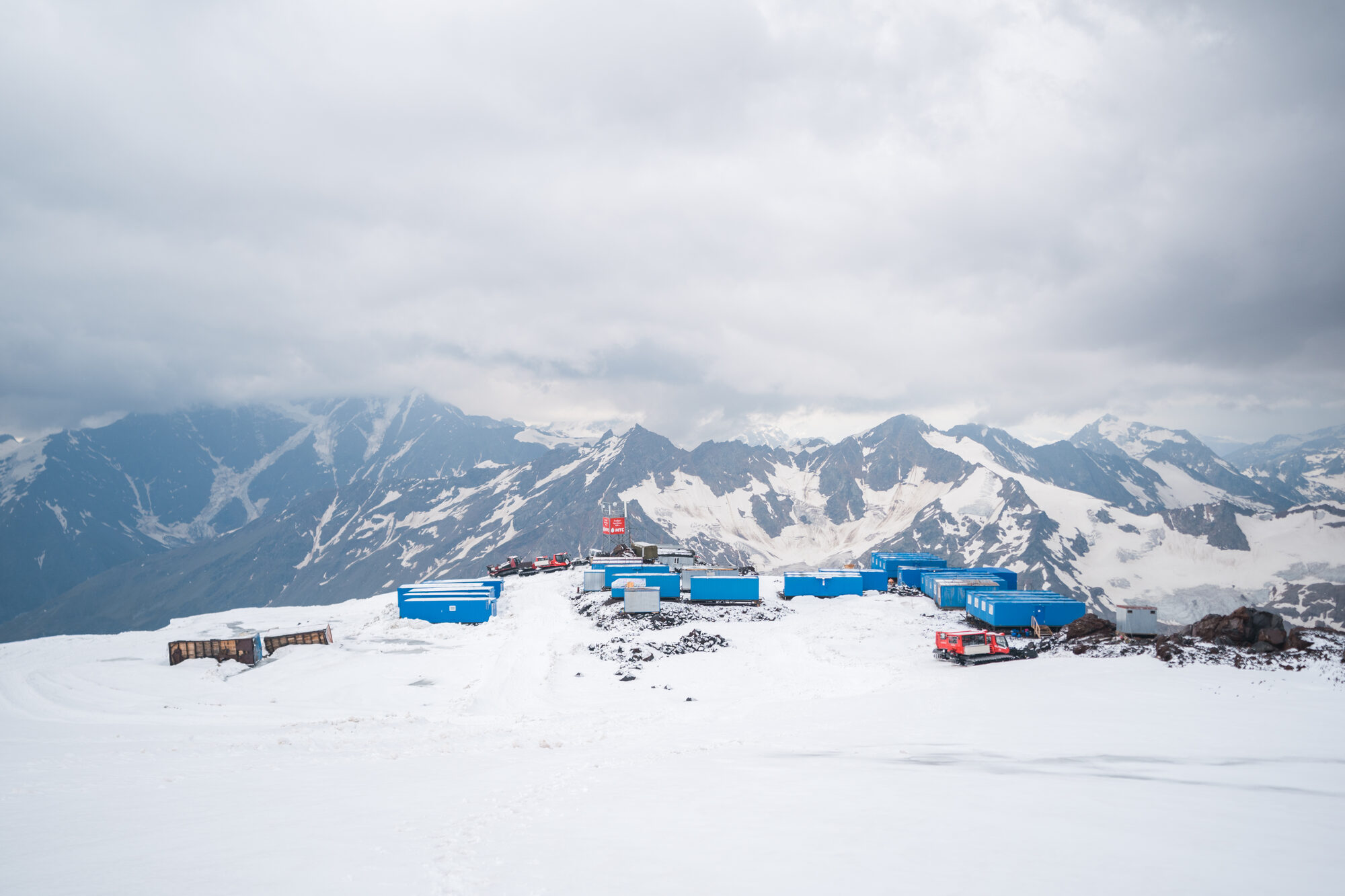 Base camp's blue container huts on the snow with the Caucasus range stretching behind